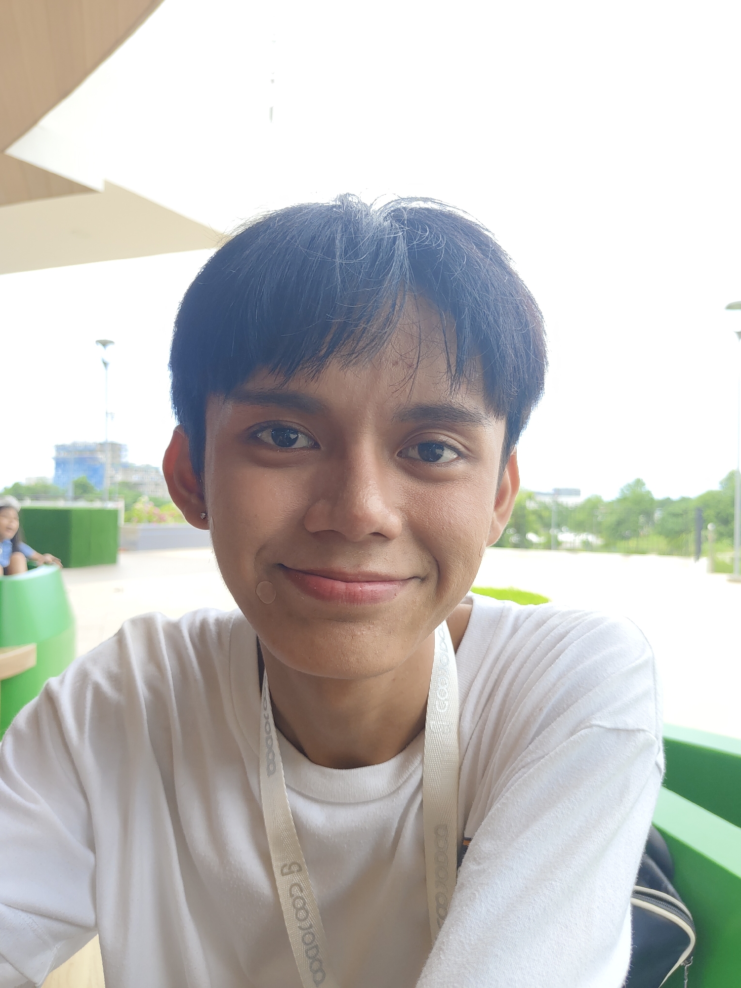 The image is a close-up, eye-level shot of a young person with dark hair, looking directly at the camera. The person is wearing a white t-shirt and a white lanyard with a logo that appears to say "EcoJobo". A small circular bandage is visible on the person's cheek. The background is blurred, showing an outdoor setting with some greenery, a building under construction, and a bright sky, suggesting a sunny day. The presence of a lanyard and the general environment are consistent with locations in Davao City, Philippines, potentially a park, a public space, or an event venue.