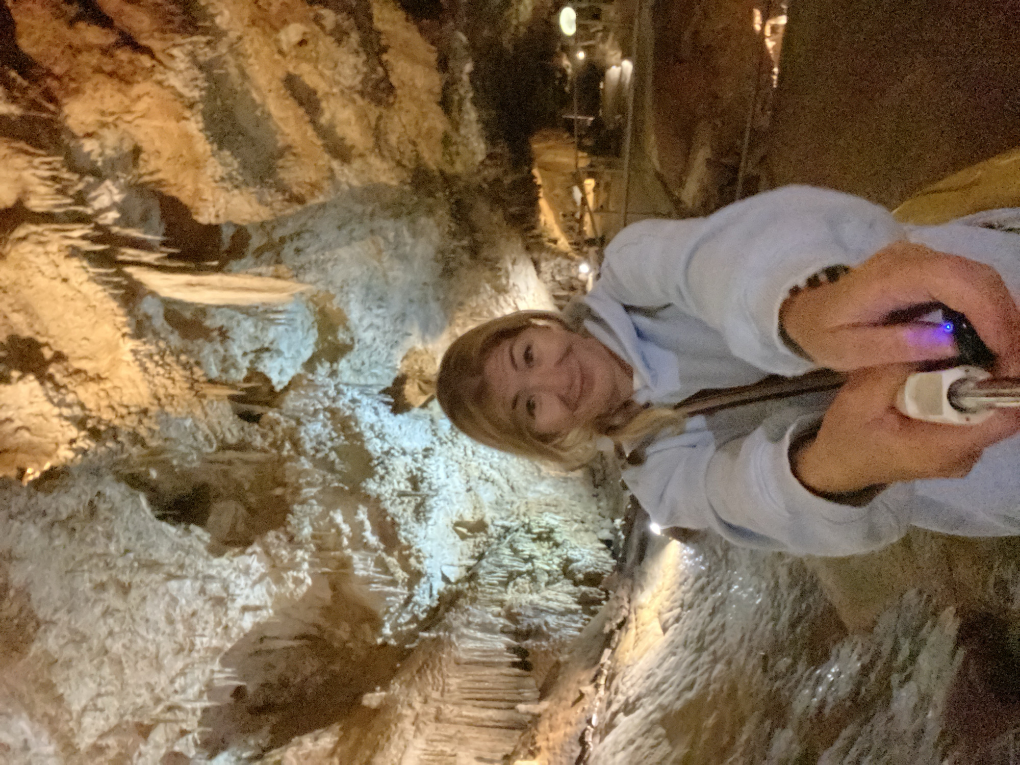 A female is taking a selfie with a cell phone inside a cave. The cave walls are textured with mineral deposits and illuminated with artificial lighting. The location is Lipa Dobrska, Montenegro.