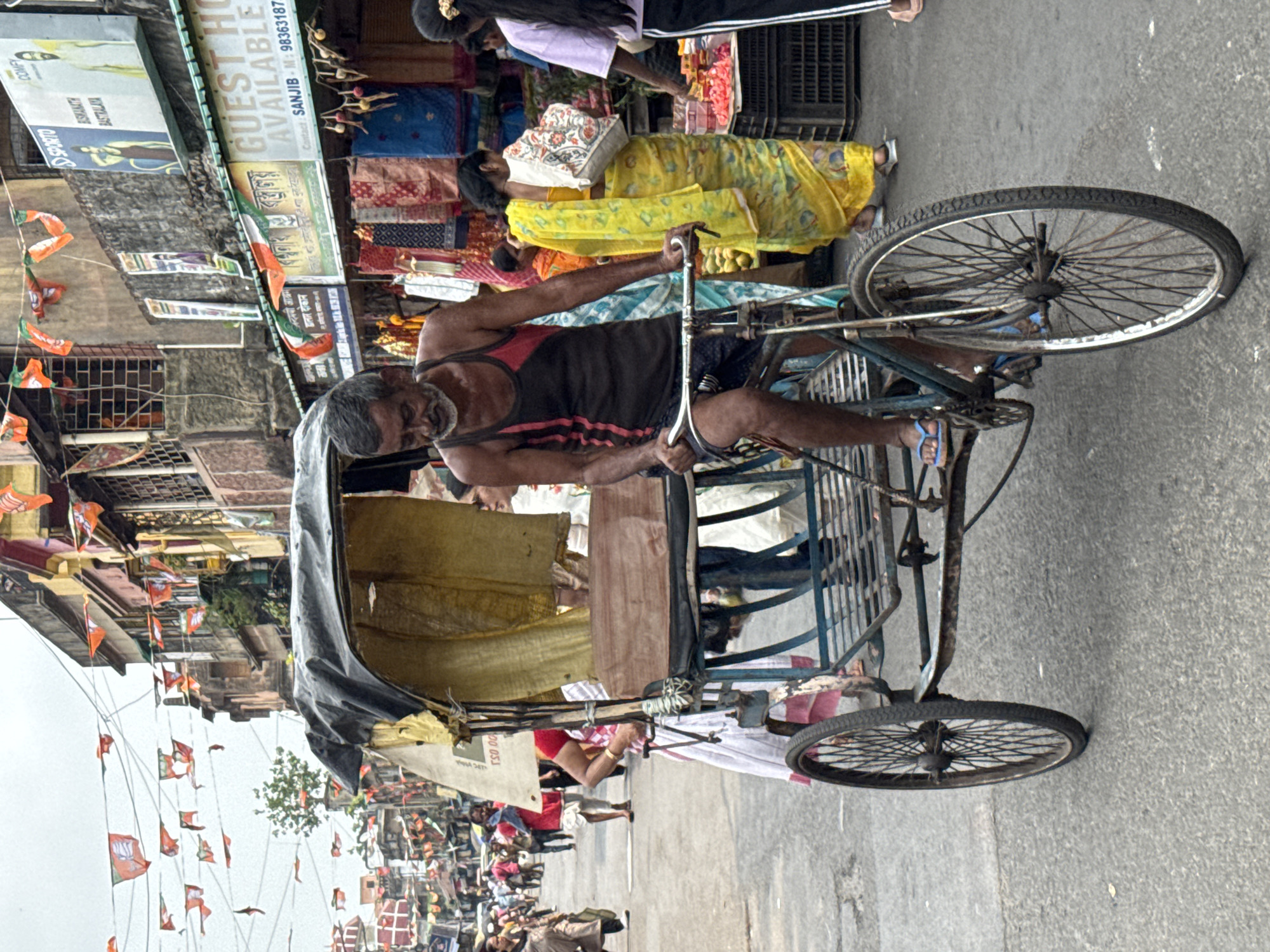 The image depicts a man riding a cycle rickshaw on a city street in Kolkata, India. He is wearing a tank top and shorts. There are passengers in the back. The street is lined with shops. BJP flags and banners are strung along the street, indicating a political event or atmosphere. Signage and storefronts display details like store names (e.g., "Biswanath Bastralaya"), contact information ("Sanjib, M: 98363187"), and phrases like "Guest House Available." Pedestrians and other vehicles are visible in the background.