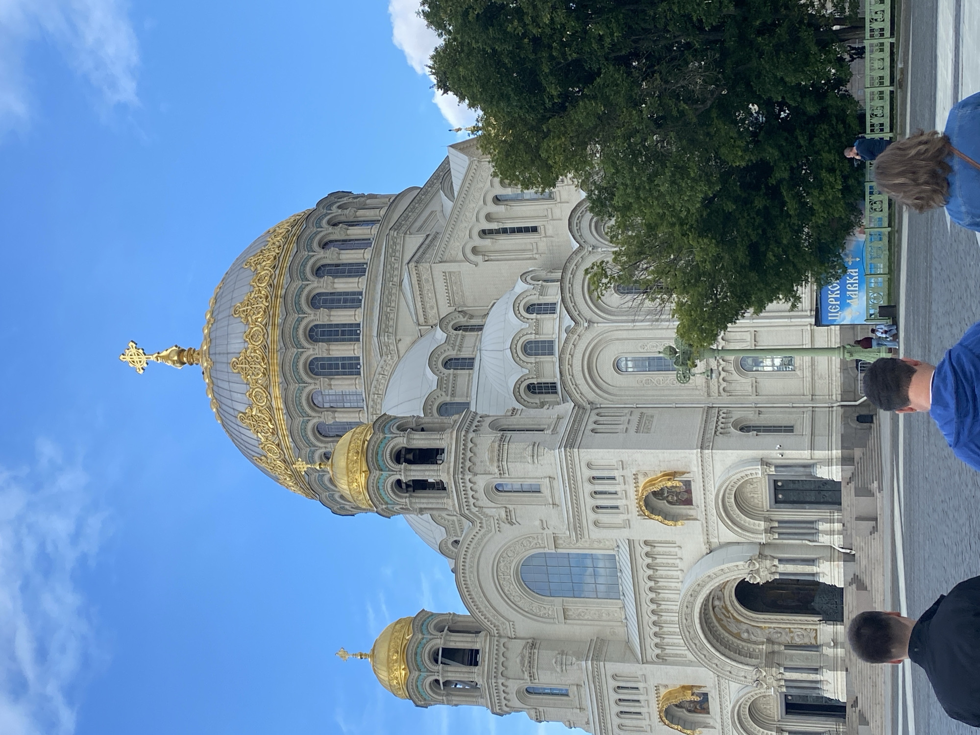 This is a medium shot, eye-level, outdoor photograph of the Kronstadt Naval Cathedral in Kronstadt, Russia, taken on a clear day with a blue sky and scattered white clouds. The grand, white cathedral dominates the frame, featuring multiple golden domes, including a large central dome topped with a cross, and two smaller domed bell towers on either side of the main entrance. The facade is adorned with arched windows and intricate architectural details, with golden accents above some of the arched doorways.
In the foreground, the backs of three individuals are visible. The person on the far left wears a black jacket. The person in the middle wears a blue hoodie. The person on the right wears a blue denim jacket. They are standing on a paved, grey surface, likely a plaza, in front of the cathedral.
To the right of the cathedral, a large, dark green tree partially obscures the building. Behind the tree, a light blue banner with Russian text is visible, along with a green decorative fence. A few other people are seen in the background, near the banner and fence, including a man talking on a mobile phone. The overall scene depicts a tourist or visitor's perspective of the landmark.