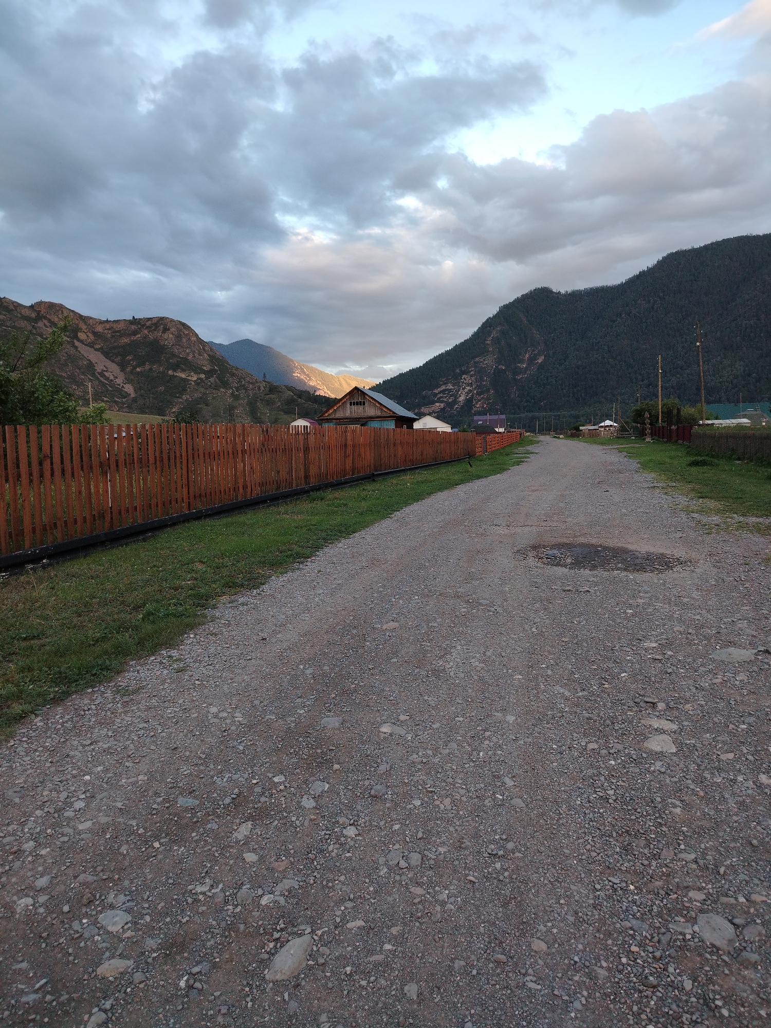 An eye-level, slightly low-angle, full shot captures a dirt road curving into the distance through a rural village in Iodro, Russia. The road is covered in gravel and has a small puddle. To the left of the road is a tall, wooden fence made of vertical planks, separating it from a strip of green grass. Beyond the fence, a residential building with a wooden exterior and a gabled roof is visible. The landscape is framed by steep, rocky mountains on the left and a dark, forested mountain on the right. The sky is overcast with clouds, but there are patches of sunlight breaking through, illuminating the peaks of the distant mountains. Power lines are visible on the right side of the road, suggesting the presence of infrastructure. The scene appears to be set in the late afternoon or early evening, given the lighting conditions. There are no people or animals visible in the image.