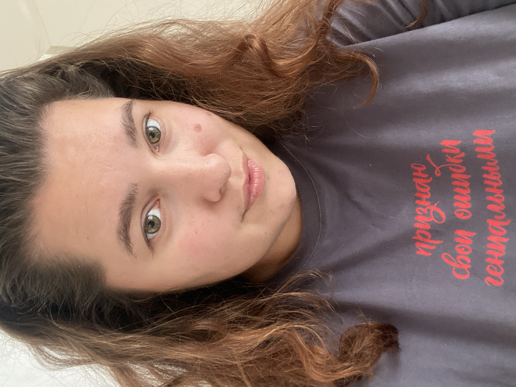A woman with auburn, wavy hair is looking at the camera. She is wearing a dark gray t-shirt with red text that reads "признаю свои ошибки гениальными". The text is in a cursive font. She has light skin with freckles and a mole on her right cheek. Her eyes are green. The background is a plain, white wall. There is no identifiable location in the city of Perm, Russia within this image.