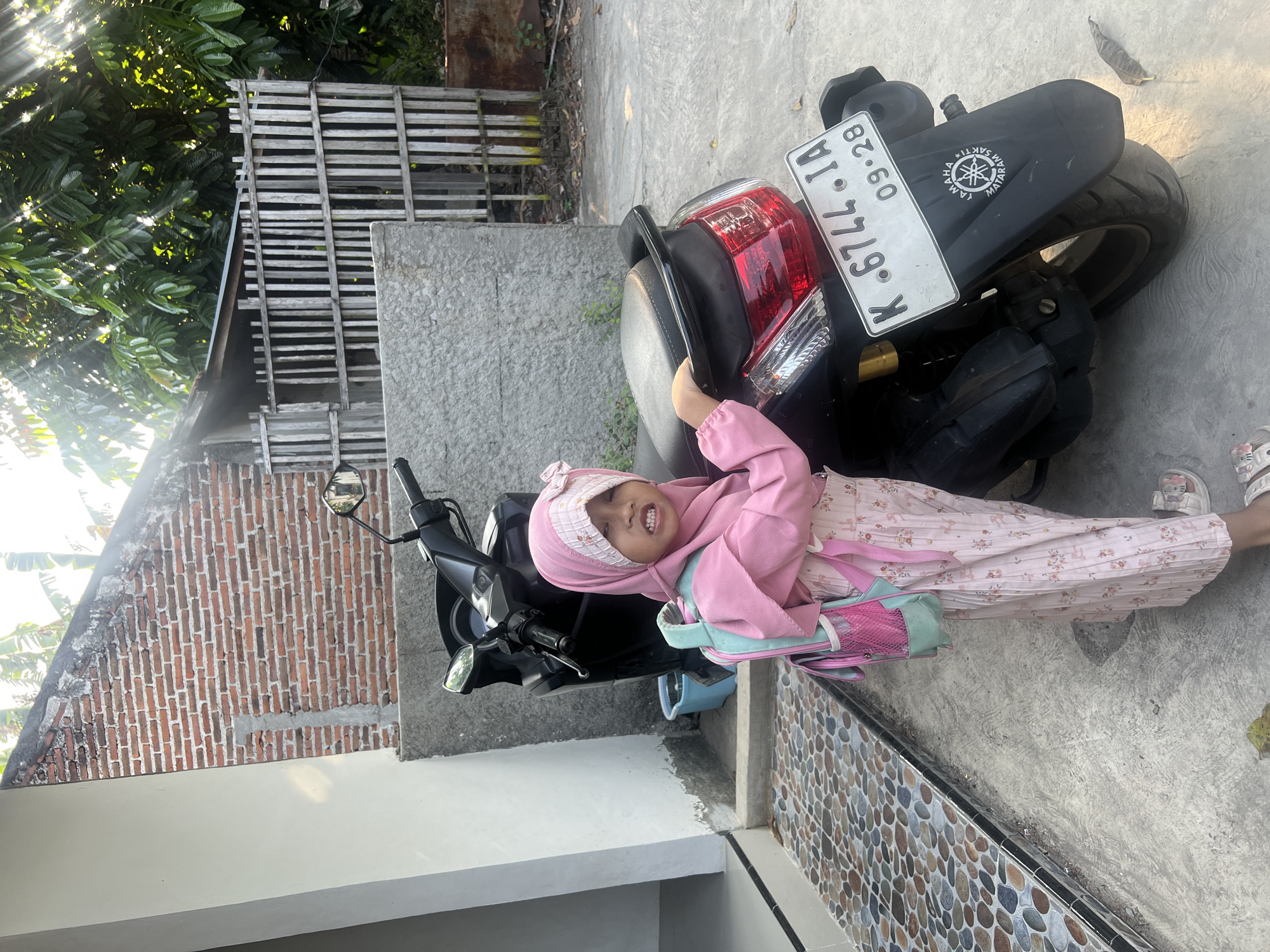 A young girl wearing a pink hijab and pink patterned clothing stands next to a black motorcycle. She has a pink and light blue backpack on her shoulders and is holding onto the back of the motorcycle. The girl is looking at the camera with a slightly open mouth, showing her teeth. The motorcycle has a white license plate that reads "K. 6744. IA" and "09-28". A "YAMAHA" logo is visible on the rear fender. The background consists of a brick wall, a concrete wall, a wooden fence, and lush green trees. Sunlight filters through the trees, creating dappled shadows on the ground. The scene appears to be an outdoor area in a residential setting, possibly in Kecamatan Pati, Indonesia, given the style of the motorcycle and the general environment. The ground is paved with small, colorful stones near a doorway on the left and concrete elsewhere.