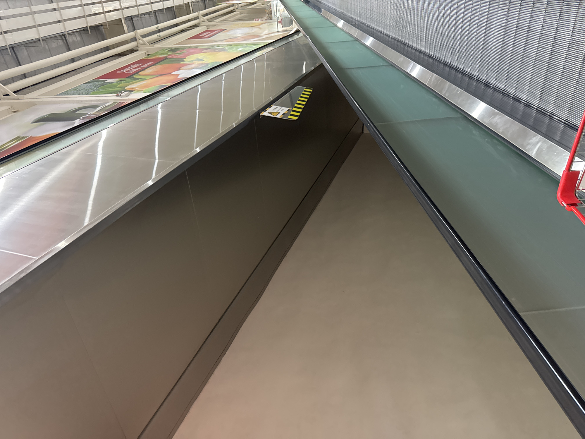 The image captures an upward-sloping moving walkway or escalator inside a brightly lit commercial establishment, likely a supermarket or large store in Bangkok, Thailand. The perspective is low-angle, looking along the length of the walkway as it ascends.
The central subject is the moving walkway, which features a grooved, metallic surface for standing. It is bordered by transparent, slightly green-tinted glass panels on both sides, topped with shiny, reflective metal handrails or trim. The left side of the walkway, from this vantage point, runs alongside a wall adorned with large, colorful advertisements. These ads primarily depict fresh produce such as fruits, vegetables, and what appears to be a rotisserie chicken, suggesting a grocery or food section. Visible Thai text on these ads includes phrases like "ยอดเยี่ยม" (Yod Yiam, meaning excellent/superb) and "สดใหม่" (Sod Mai, meaning fresh).
Mid-way up on the left side, a small, triangular warning sign with black and yellow diagonal stripes and a caution symbol is affixed to the side of the moving walkway, indicating a safety alert. To the far right, a small portion of a red shopping cart is visible, further reinforcing the retail setting. The area beneath the moving walkway is an empty, light-colored space. The ceiling structure, with its white beams and pipes, is partially visible in the upper background. The bright artificial lighting within the store creates strong reflections on the polished metal and glass surfaces, enhancing the modern and clean aesthetic of the space. No people are visible in the frame, giving the impression of an early morning or late evening scene, or simply a moment without heavy foot traffic.