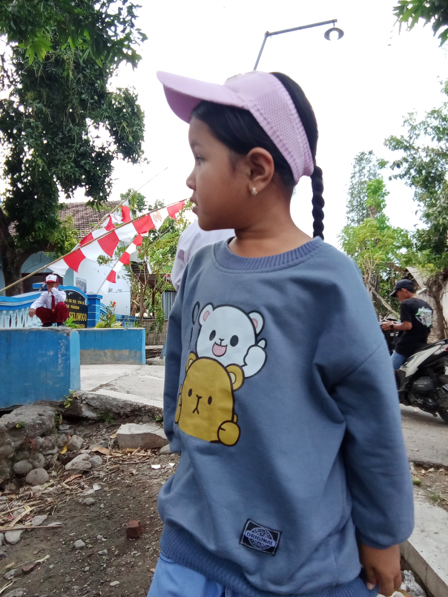 A young girl, viewed from the side, stands in an outdoor setting. She has dark hair braided into a single plait and wears a light pink visor. Her grey sweatshirt features a graphic of two cartoon bears. She is looking to her left, away from the camera. In the background, a person in a red and white uniform is crouching near a blue structure with the sign "SELOWOGO". Red and white triangular flags are strung across the scene. To the right of the girl, a person on a motorcycle is visible. The ground is covered in dirt, rocks, and dry leaves. The scene appears to be in a semi-urban or village environment in Trebungan, Indonesia, suggested by the architecture and flags, which might indicate a national holiday or celebration.