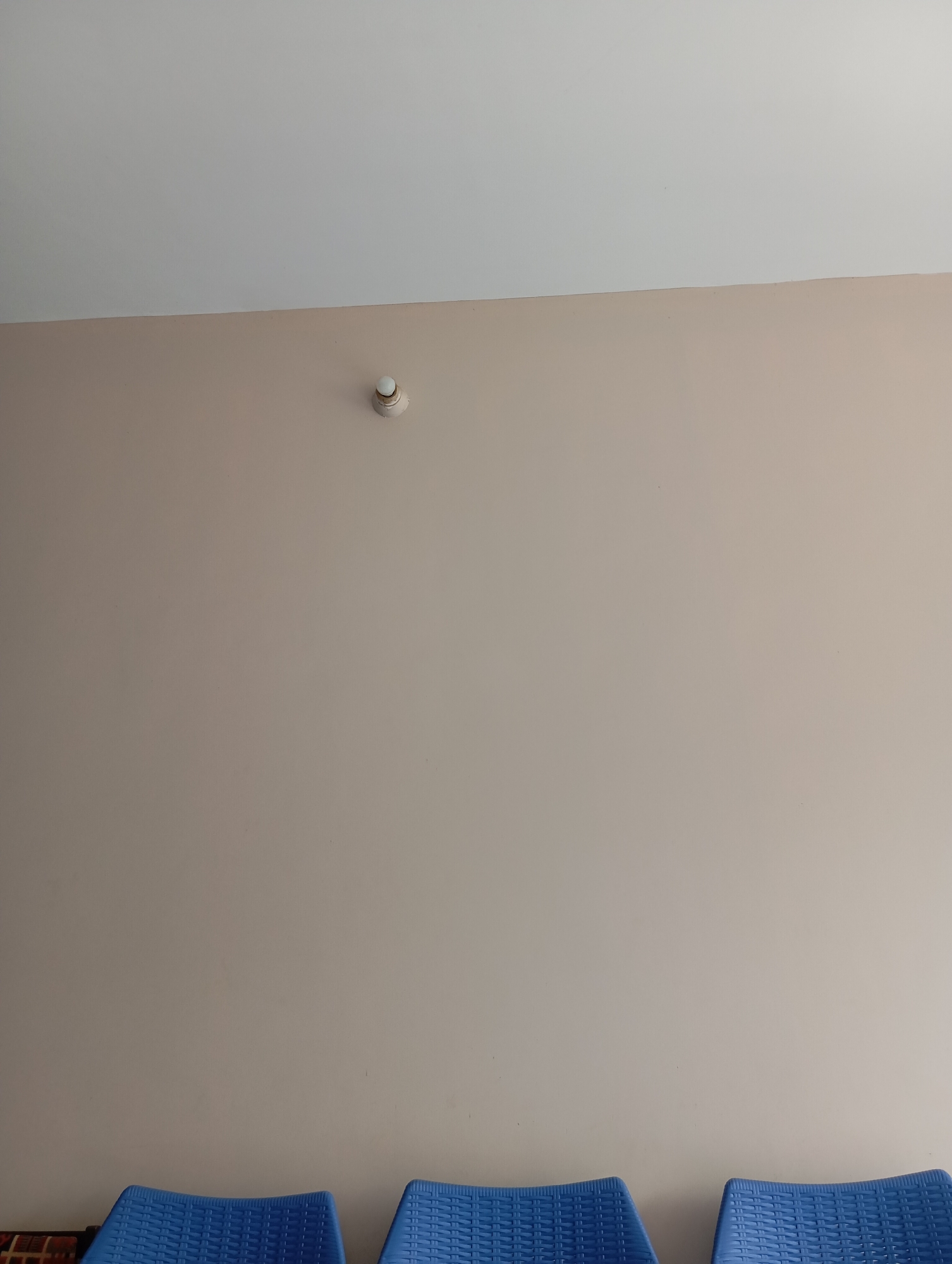 A smoke detector is mounted on a light-colored wall in an indoor environment, possibly a waiting room or office. The wall is painted a neutral tone and is adjacent to a white ceiling. In the foreground, the tops of three blue plastic chairs are visible, suggesting a seating area. The scene is static and does not depict any ongoing activity or individuals.