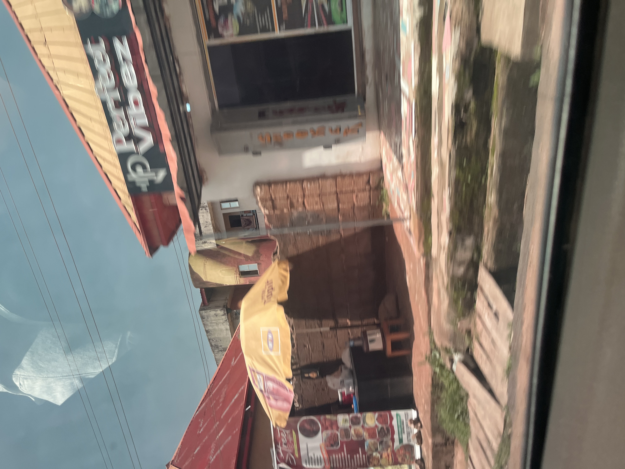 The image shows a street scene in Jos, Nigeria. In the foreground, a wooden walkway crosses a drainage ditch. Behind the walkway, a food stall operates under a yellow umbrella. A menu board displays food items. A building with red roofing is visible behind the food stall. Above the food stall, a sign reads "Perfect Vibez." The sky is visible in the background. The scene appears to be a typical roadside establishment in a Nigerian city.