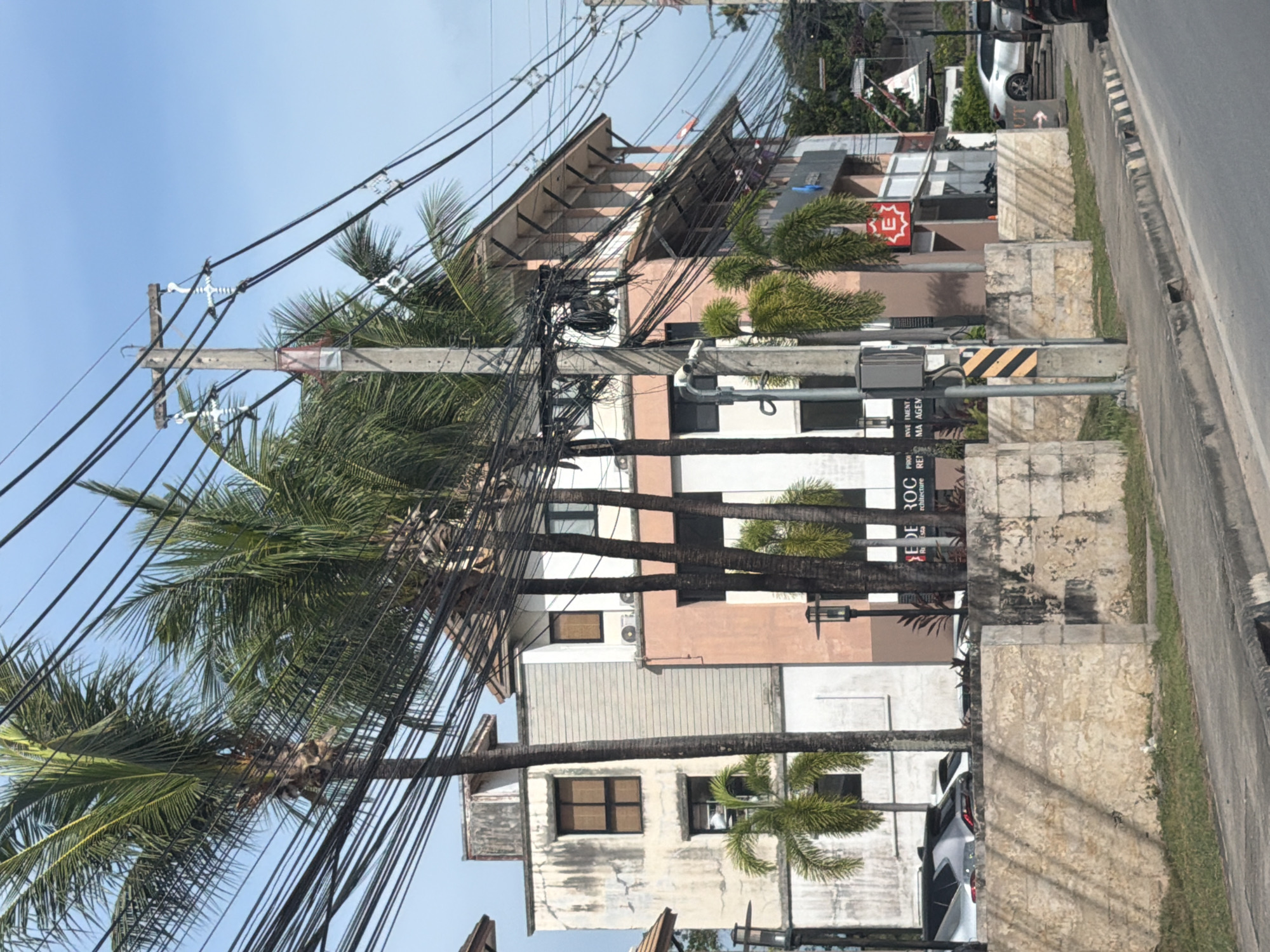 The image shows a street scene in Baan Bo Phut, Thailand, on a clear day. The foreground features a concrete sidewalk and curb, with a grassy area behind it. A low, rough-hewn stone wall runs parallel to the sidewalk. In the midground, a multi-story building with a peach-colored facade and white accents is visible, adorned with palm trees and electrical wires. Several windows and air conditioning units are present on the building. To the left, a lighter-colored building with a weathered facade and dark-paned windows stands. The background is dominated by a bright blue sky and a dense network of black electrical wires strung between utility poles. A sign with red lettering and a white 'E' inside a red square is visible on the peach-colored building, possibly indicating a business. Cars are parked on the street to the left, and a white car is seen driving on the right side of the road. The scene suggests a commercial or residential area with a tropical influence due to the palm trees.