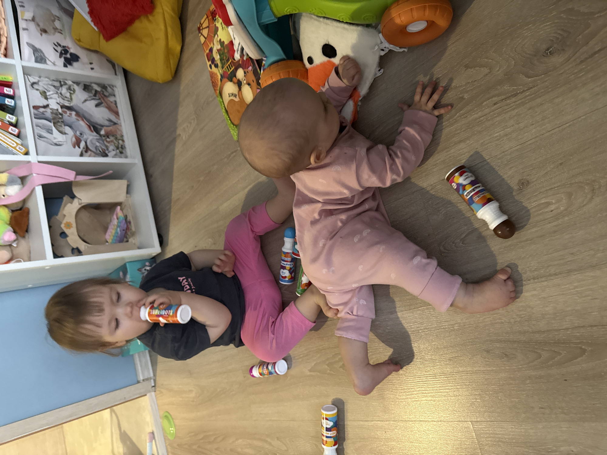 Two infants are lying on a light-colored wooden floor surrounded by toys. One infant is wearing a pink onesie and the other is wearing a dark grey t-shirt and pink pants. Both children are holding tubes of paint. There are multiple paint tubes scattered on the floor around them. A white shelf with toys and storage boxes is visible in the background. A colorful play mat is also present. The location appears to be inside a home or daycare, potentially in Moscow, Russia.