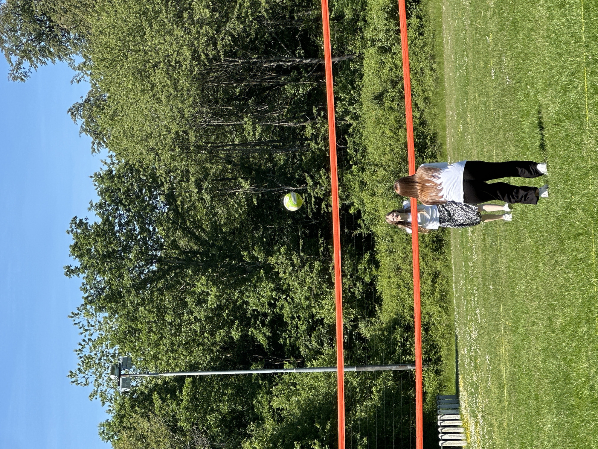The image shows a scene in Grünberg, Germany. Two girls are standing on a grassy field with dandelions. One is wearing a white t-shirt and black pants, the other a white t-shirt and a black and white patterned skirt. There is an orange goalpost visible. A green and white ball is suspended in the air, seemingly between the goalposts. Trees and a blue sky are in the background. A metal pole with lights is also visible behind the trees. A wire fence is situated behind the orange goalposts.