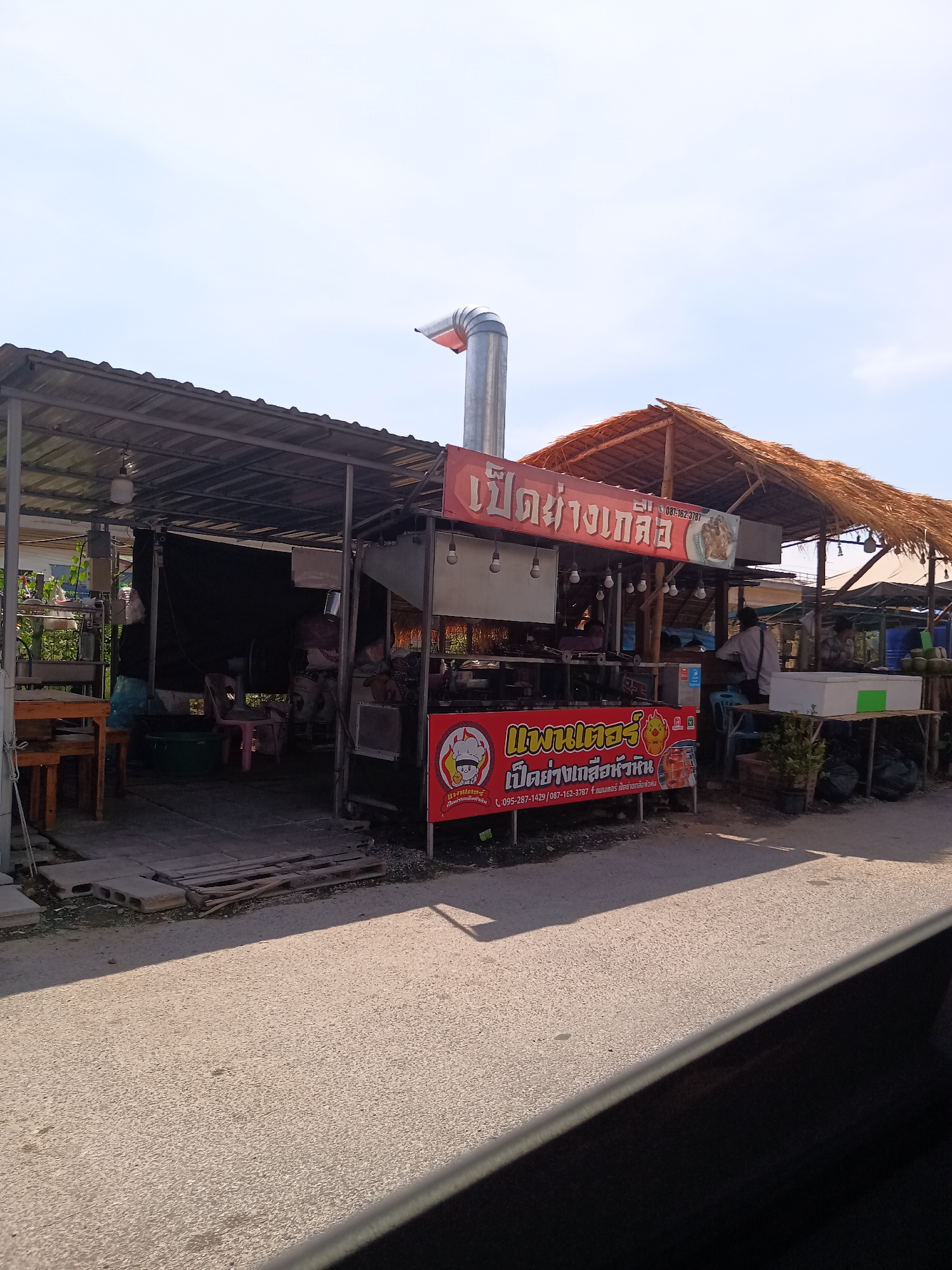 The image shows a street-side food stall in Hua Hin City Municipality, Thailand. The stall is partially covered with corrugated metal roofing and features a thatched roof section. Prominent signage in Thai script advertises "Pet Yang Kleu," (grilled salty duck) and includes a phone number. The stall is equipped with a tall metal exhaust pipe. People can be seen inside the stall. The street is paved.