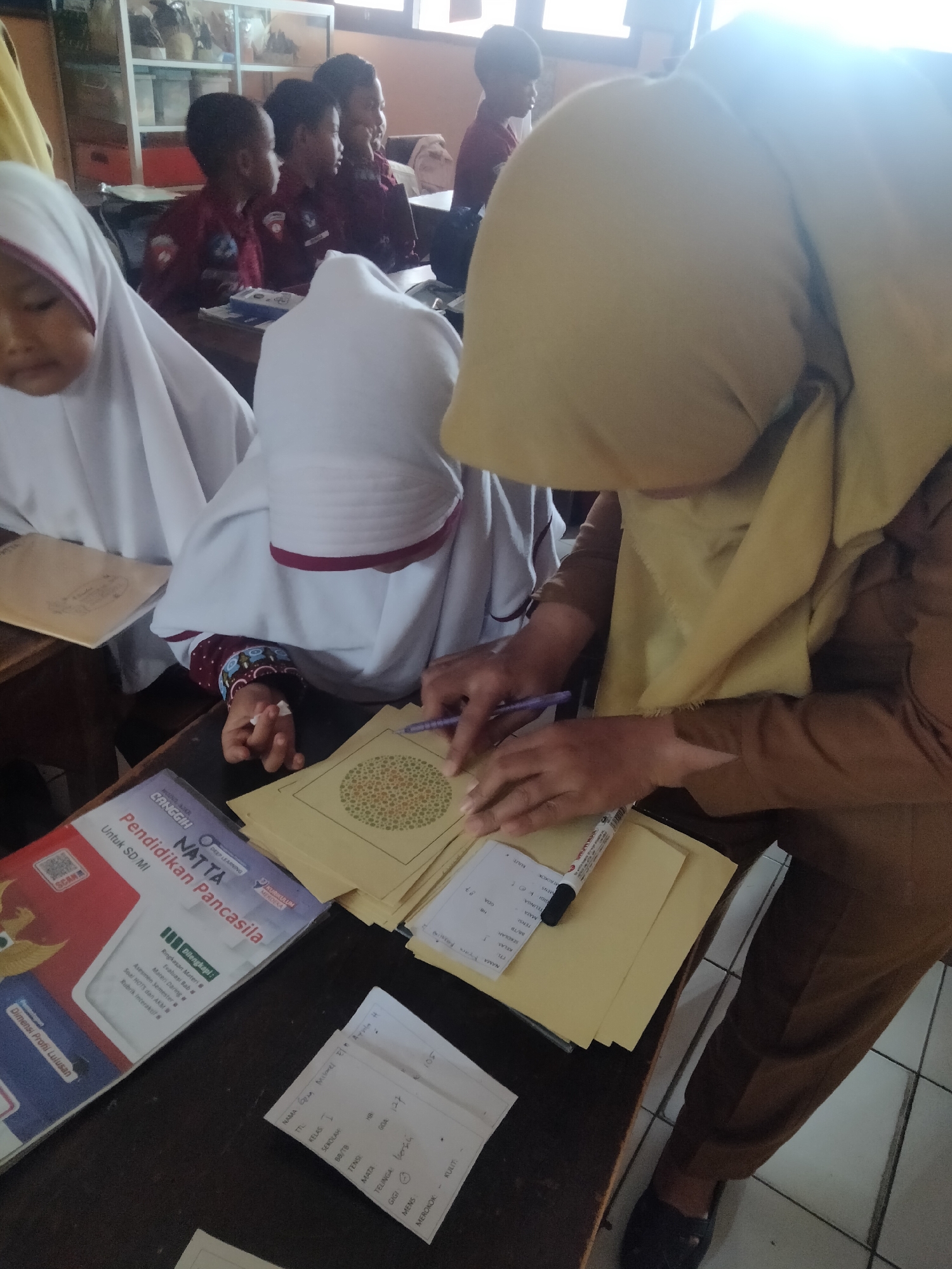 In a classroom setting in Tretes, Indonesia, a group of children are participating in an eye test. The image focuses on a student wearing a white hijab, her left hand holding a pen over a color blindness test chart. Beside her, another student in a white hijab is also visible, with their face partially obscured. In the background, several other children are seated, some wearing maroon uniforms. A teacher or adult, identifiable by a brown uniform and a yellow hijab, is also present, assisting with the test. The room appears to be a typical school environment, with desks, chairs, and educational materials visible. The overall scene suggests a routine medical or educational screening taking place.