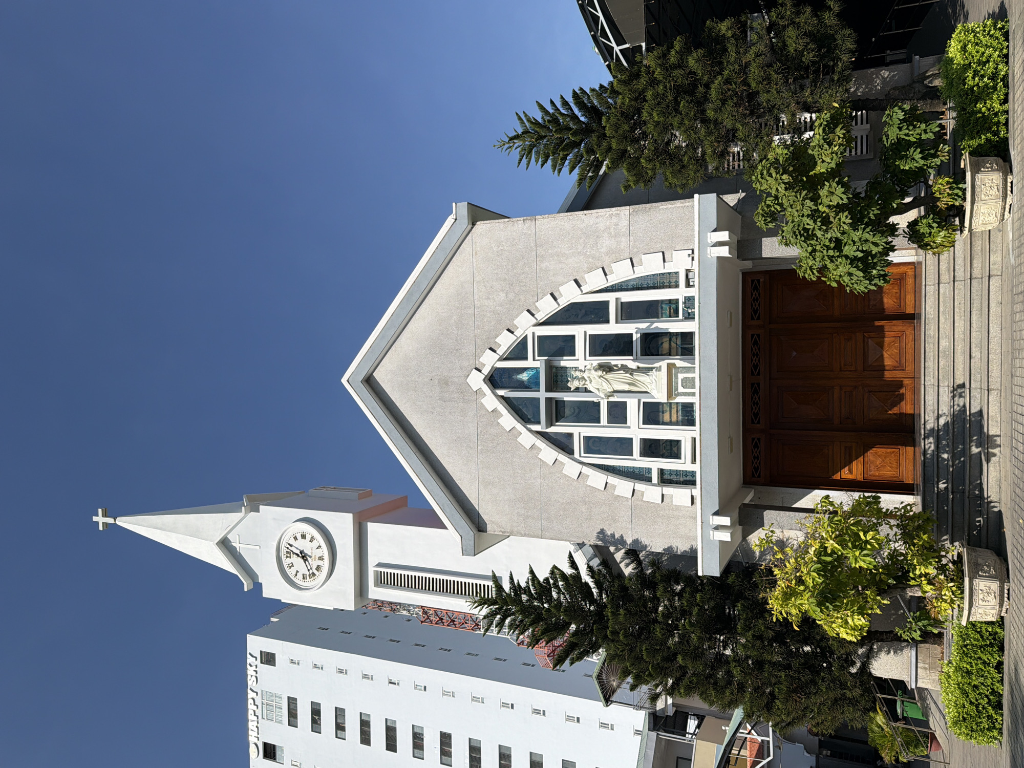The image shows the façade of a church with a tall, white bell tower topped with a cross, located in Nha Trang, Vietnam. To the left of the church, a tall, modern white building with many windows is visible, bearing the "HAPPY SKY" signage. The church itself has a grey, textured exterior, with a prominent arched window featuring stained glass and a statue of a person, likely a religious figure, above the entrance. The entrance features large, wooden double doors and is accessed by a set of stairs. Two evergreen trees flank the church entrance, with a potted plant in the foreground and a manicured hedge to the left. The sky is clear and blue, indicating a sunny day. The scene appears to be a street view of the church and adjacent buildings.