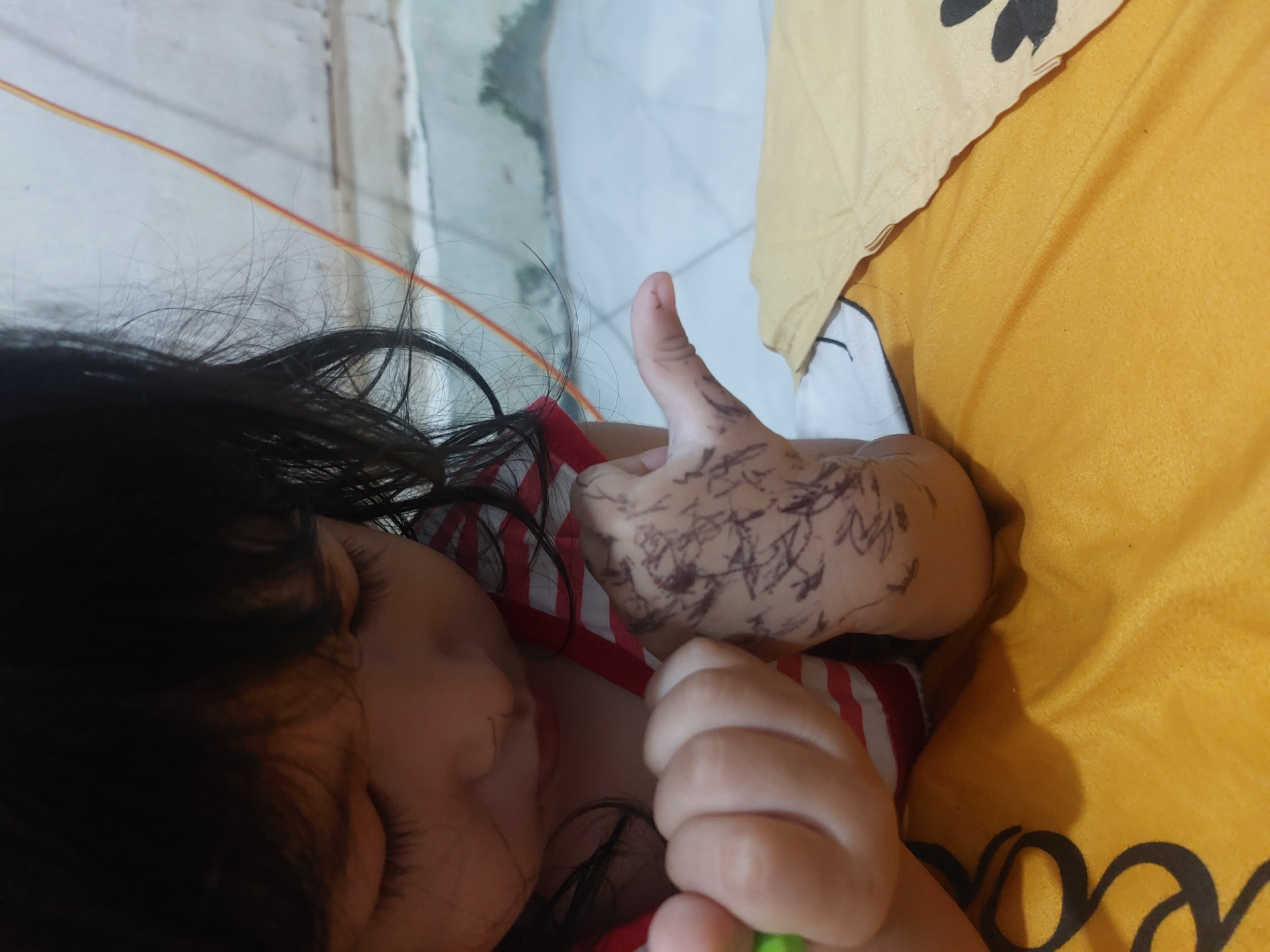 The image captures a close-up, top-down view of a young child engaged in a playful activity indoors, likely in Lhokseumawe, Indonesia. The child, with dark hair, is partially visible, lying down or resting on a surface.
The central focus is the child's right hand (viewer's left), which is covered in scribbled drawings made with a dark marker or pen, showcasing a creative act common among children. This hand is held up, displaying a "thumbs-up" gesture. The child's left hand is also visible, clenched into a fist in the lower right foreground, with a small green tip, possibly from the marker used for the drawings, barely discernible. The child is wearing a red and white striped top.
The setting appears to be an indoor space. A large yellow-orange fabric, possibly a blanket or bedspread, covers a significant portion of the scene. Visible on this fabric is a dark, partial pattern or text that resembles "coco". Above this, a lighter yellow-beige fabric is also present. The background shows a light-colored, possibly tiled, floor or wall. The child's face is mostly obscured by their dark hair, but the visible portion of the cheek and mouth suggests a relaxed and absorbed state during the activity. The overall scene depicts a moment of innocent, creative play.