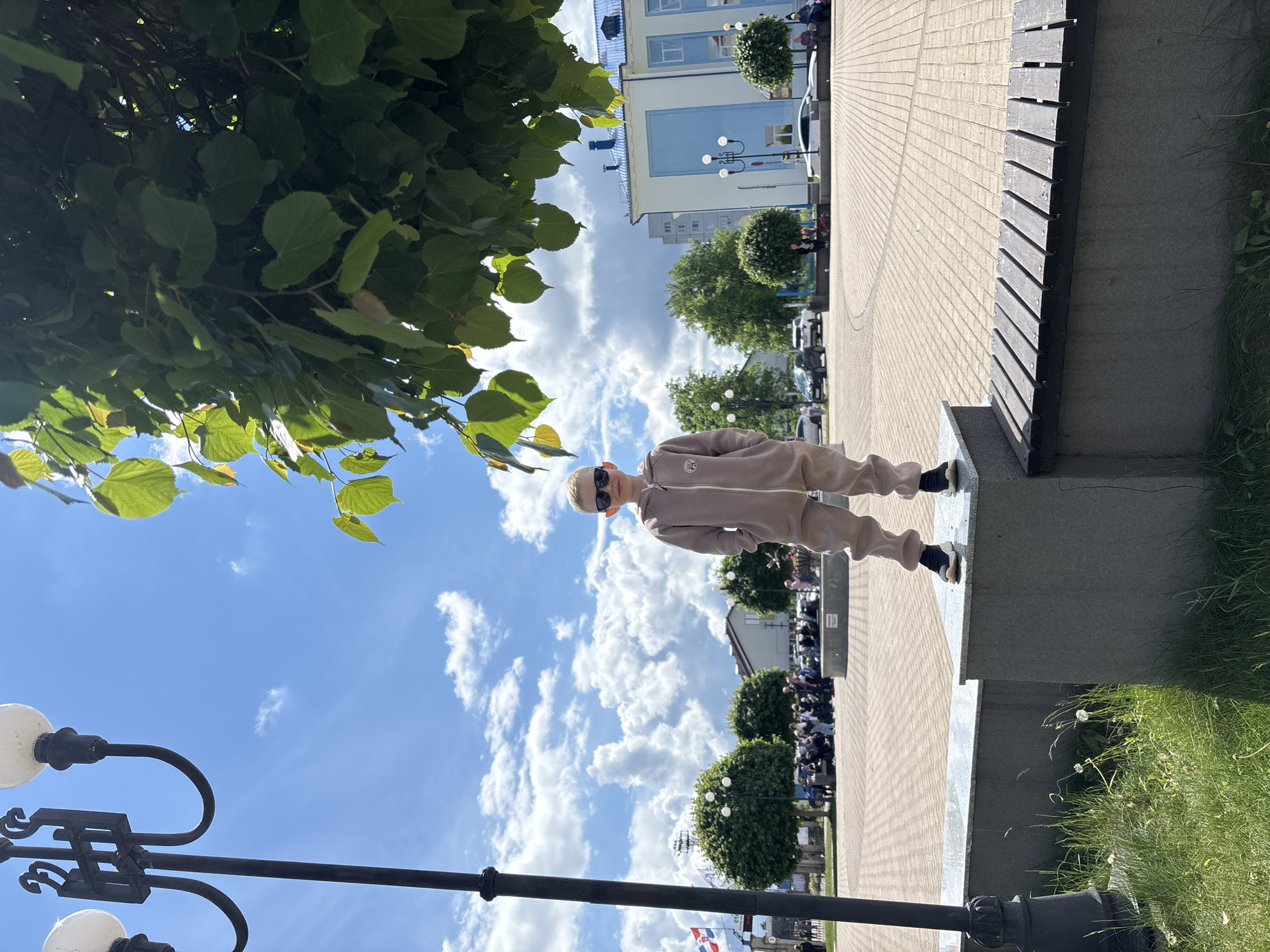 A young person stands on a concrete ledge beside a wooden bench in the city of Chaikovsky, Russia. The person wears a tan tracksuit, black sneakers, and sunglasses. The background shows a brick-paved plaza with decorative spherical streetlights, green trees, and a building with blue and white elements. In the distance, a crowd of people is visible. A lamppost is partially visible on the bottom left, and tree foliage partially obscures the top left of the frame. The sky is visible, with fluffy clouds.