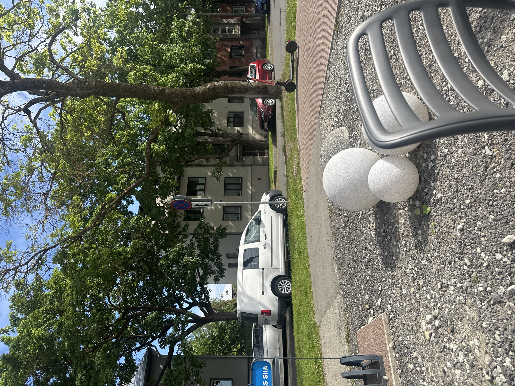 The image depicts a sunny day in an urban setting, likely in Tallinn, Estonia, given the presence of a blue sign with white text that reads "AS SIIA," which is a known supermarket chain in Estonia. In the foreground, a dark gray plastic chair is partially visible, facing away from the camera. To the left of the chair, three large, speckled gray spheres are arranged on a bed of gravel. A metal bicycle rack is also present in this area, adjacent to a reddish-brown paved section.
The middle ground features a paved pathway and a grassy verge. Several vehicles are parked along the street, including a white van, a white passenger car, and a red SUV. An electric scooter is parked on the pathway near the red SUV. Behind the vehicles, apartment buildings with multiple windows and balconies line the street. A traffic sign with a red circle and a horizontal bar, along with a double-headed arrow below, is visible on a pole in front of one of the buildings.
The background is dominated by lush green trees, their leaves vibrant in the sunlight, suggesting a spring or summer season. The sky is blue with some scattered white clouds. The overall impression is a peaceful street scene in a residential area, with a mix of nature and urban infrastructure.