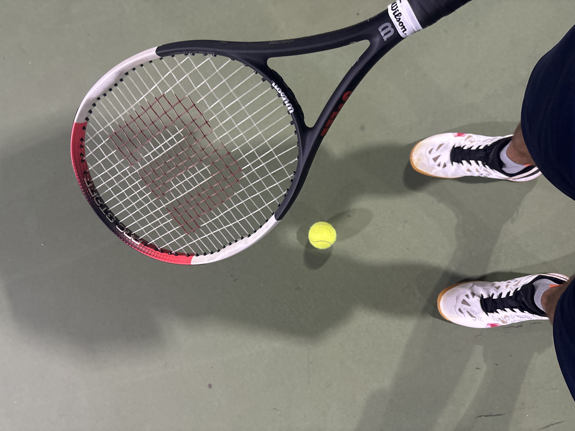 An overhead shot captures a tennis racquet and ball on a green court. The racquet, positioned diagonally from the upper right to the lower right, is black with white and red accents. A bright yellow tennis ball rests on the court to the left of the racquet. In the lower portion of the image, the feet of a person wearing white tennis shoes with black laces are visible, suggesting they are standing on the court. The court surface appears to be a standard green tennis court, possibly indoors given the lighting and shadows. There are no other people or discernible city landmarks visible.
