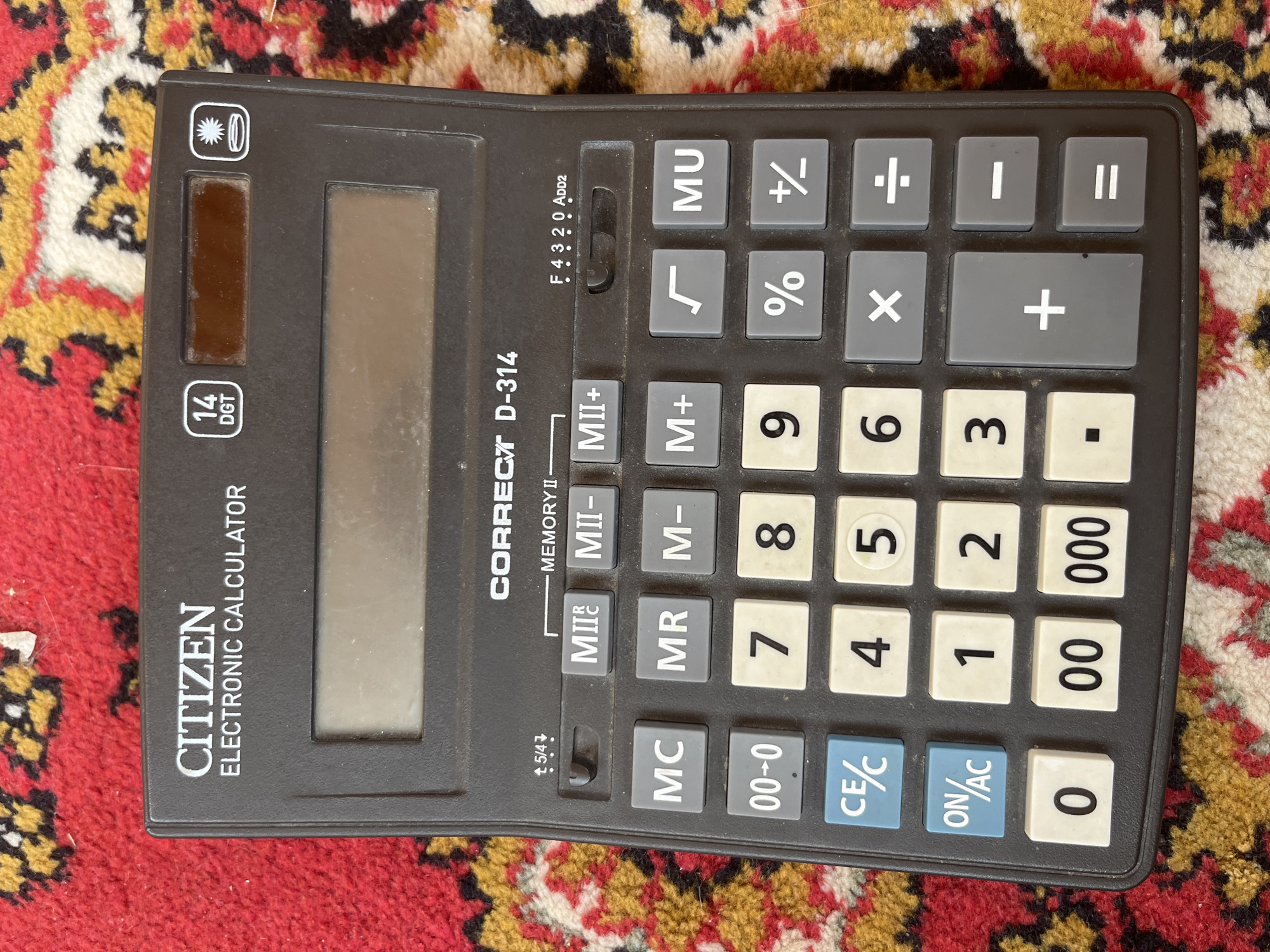 A dark grey Citizen Correct D-314 electronic calculator is positioned centrally on a multi-colored patterned rug in Tyumen, Russia. The calculator features a 14-digit display capacity, indicated by "14 DGT," and includes a solar panel element visible in the upper right. Its large, rectangular liquid crystal display (LCD) is noticeably dusty. Above the main keypad, two slider switches control calculator functions: one for rounding (marked 5/4 with directional indicators) and another for decimal point settings (F, 4, 3, 2, 0, ADD2). The keypad comprises 48 buttons; numeric keys (0-9, 00, 000, .) are white with black text, while most function keys (MC, MR, M-, M+, MII C, MII-, MII+, √, %, +/-, MU, x, ÷, -, =) are various shades of grey with white symbols. The ON/AC and CE/C buttons are light blue with white text. The calculator shows signs of moderate use and dust accumulation. The background rug displays a traditional pattern with a dominant red field interwoven with yellow, white, and dark brown/black motifs.