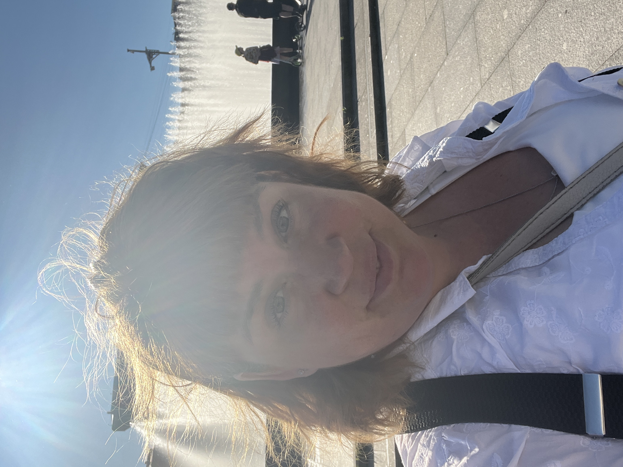 A young woman with shoulder-length brown hair takes a selfie in an outdoor setting, with the sun bright and casting lens flares across the image. She is wearing a white, embroidered blouse and a black belt. The background is slightly blurred, but appears to be a paved area with people in the distance, possibly near a fountain. The sky is clear and blue. The time of day appears to be daytime due to the bright sunlight. The overall mood seems casual and relaxed. The location is identified as Saint Petersburg, Russia.