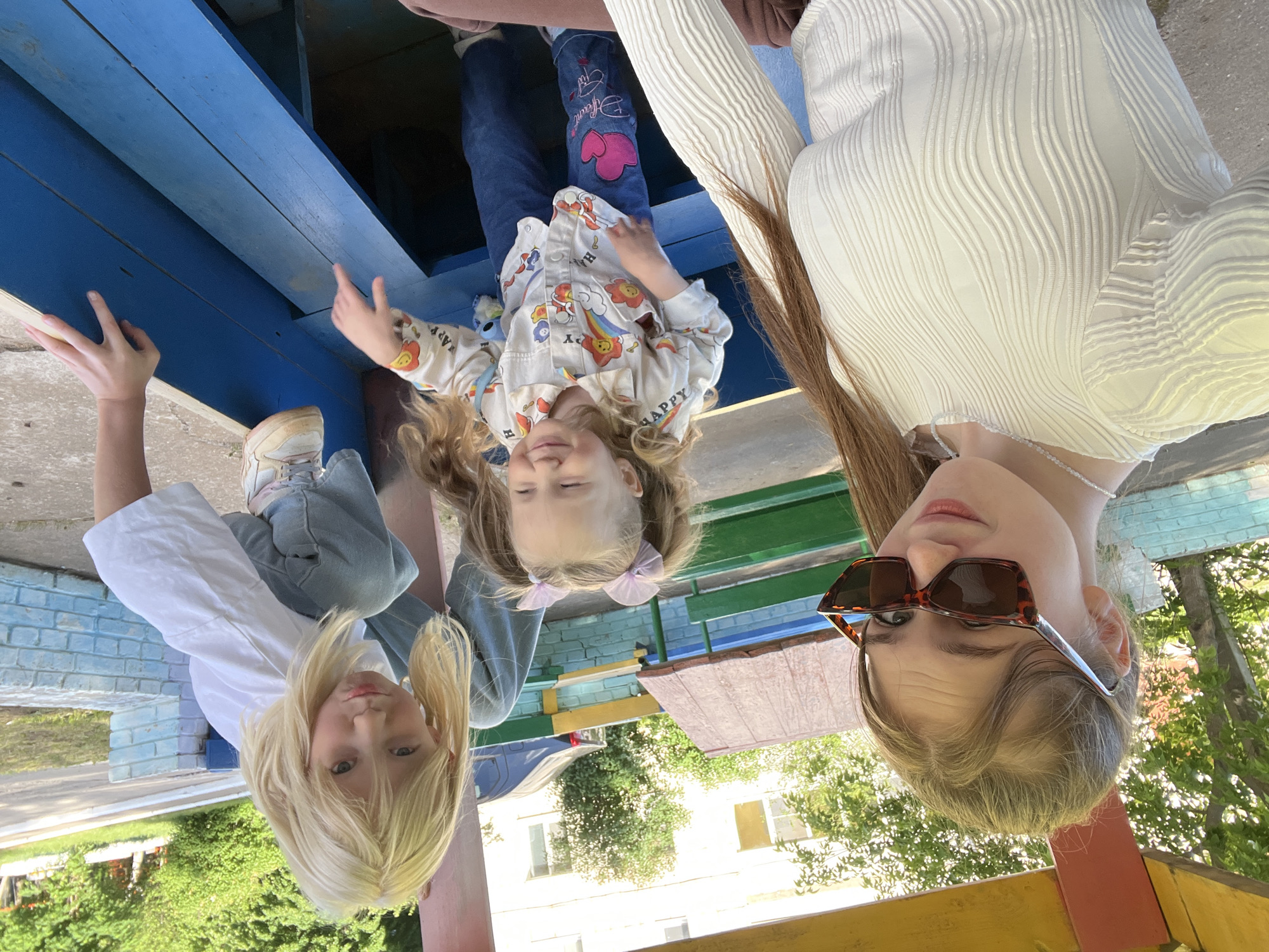Three girls are visible in the image. A young girl in a patterned shirt and jeans is in the center. She has blonde hair and is wearing purple bows. To the left is another young girl with blonde hair in a white shirt and jeans. To the right is a teenage girl with long brown hair wearing sunglasses and a white blouse. The girls are positioned in and around a blue and yellow structure, potentially a playground or outdoor play area. The background suggests an outdoor setting with trees and buildings, possibly in Chaikovsky, Russia.