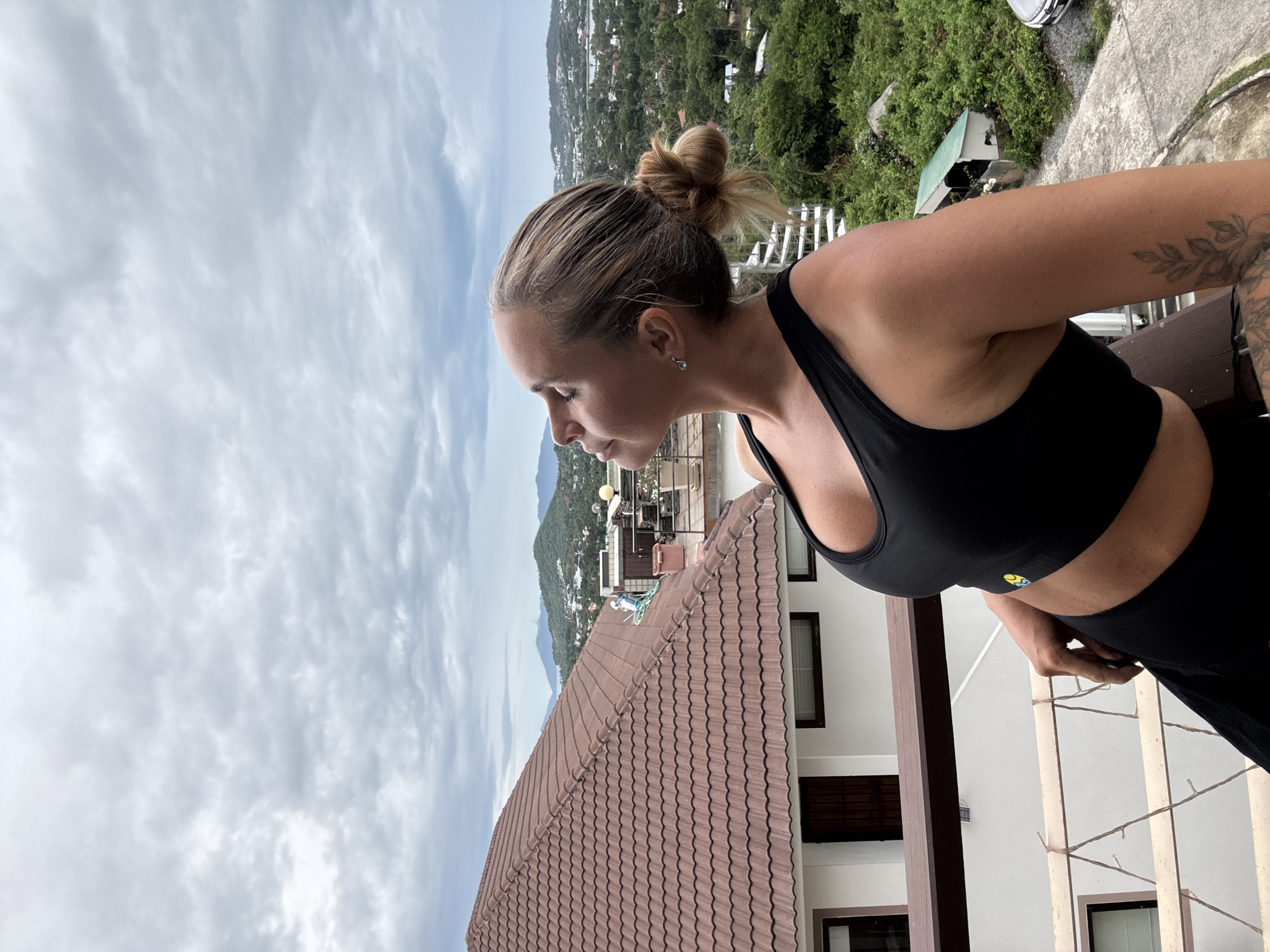 A woman with blonde hair pulled into a bun is standing on a balcony overlooking a cityscape and lush green hills under a cloudy sky. She is wearing a black sports bra and black leggings. Her head is tilted back and her eyes are closed, as if basking in the warmth of the sun. The background shows a tiled roof of a building, several other structures, and a dense green landscape with mountains in the distance. The architecture and vegetation suggest a tropical location like Ko Samui District, Thailand.