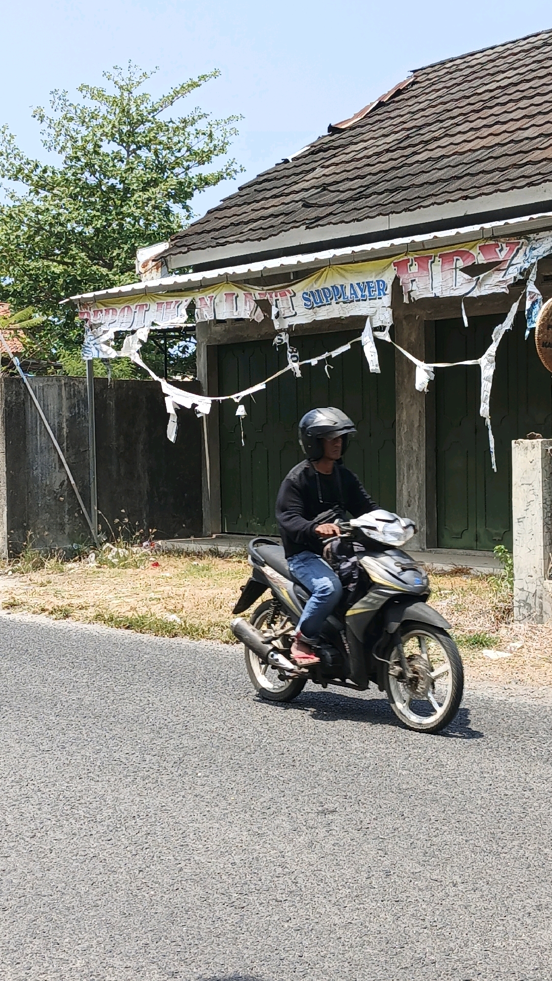 A person wearing a black long-sleeved shirt, blue jeans, red sandals, and a black helmet rides a black and yellow scooter down a paved road. The scooter has a silver exhaust pipe and white rims. Behind the scooter is a weathered building with a dark brown tiled roof and green double doors. A tattered banner with the word "SUPPLAYER" is draped across the front of the building. To the left of the building is a grey concrete wall and dry vegetation. The scene is outdoors on a clear day, suggesting a rural or semi-rural setting in Desa Karangjaladri, Indonesia.