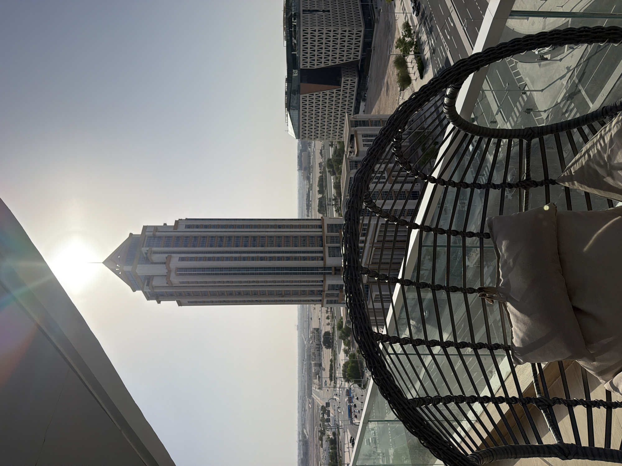 The image displays a high-angle view of a cityscape on a sunny day, captured from a balcony or elevated viewpoint. The scene is dominated by modern architecture, including a prominent, tall skyscraper with a distinctive pointed top, and another building with a geometric, lattice-like facade. In the foreground, a portion of a wicker chair with light-colored cushions is visible, suggesting a relaxed, outdoor setting. The chair is positioned to overlook the urban landscape.
The sun is high in the sky, casting bright light and lens flare across the image, indicating it is likely midday. The sky is clear and bright, suggesting fair weather. Below the buildings, a network of roads with some vehicular traffic can be seen, hinting at a bustling city environment. The overall atmosphere appears calm and serene, with a sense of urban expanse. The image is oriented vertically, with the cityscape appearing as if viewed from the side. No visible text is present in the image.