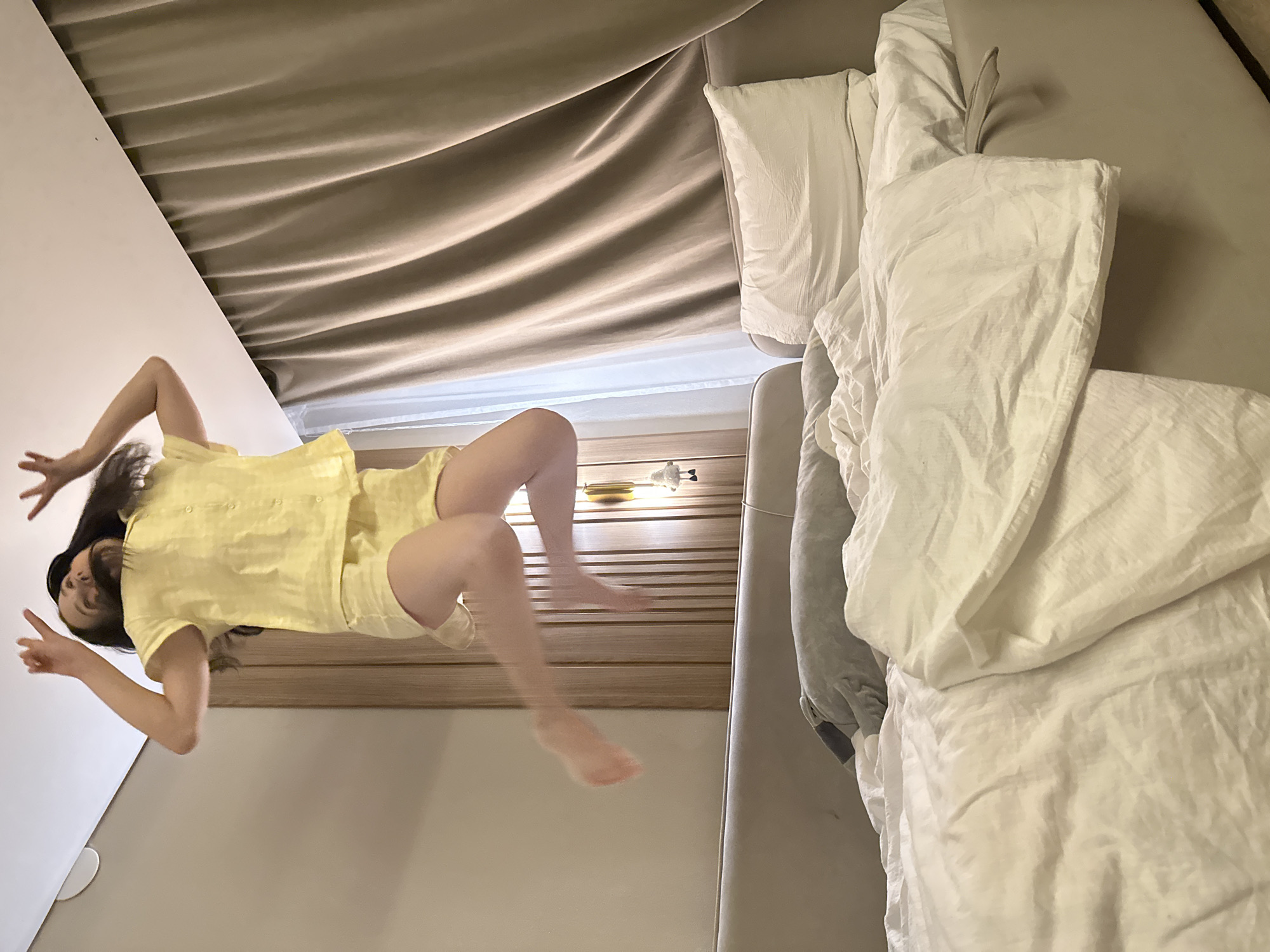 A person in a yellow short-sleeved shirt and shorts is positioned in front of a wall and a bed. The person's feet are visible in the air, indicating movement or a jump. The wall is white, and the bed has white pillows and sheets. The image includes a headboard, a wall, and what appears to be a curtain. There is no information provided that states the location is Votkinsk, Russia. Therefore, I cannot include the city in the description.