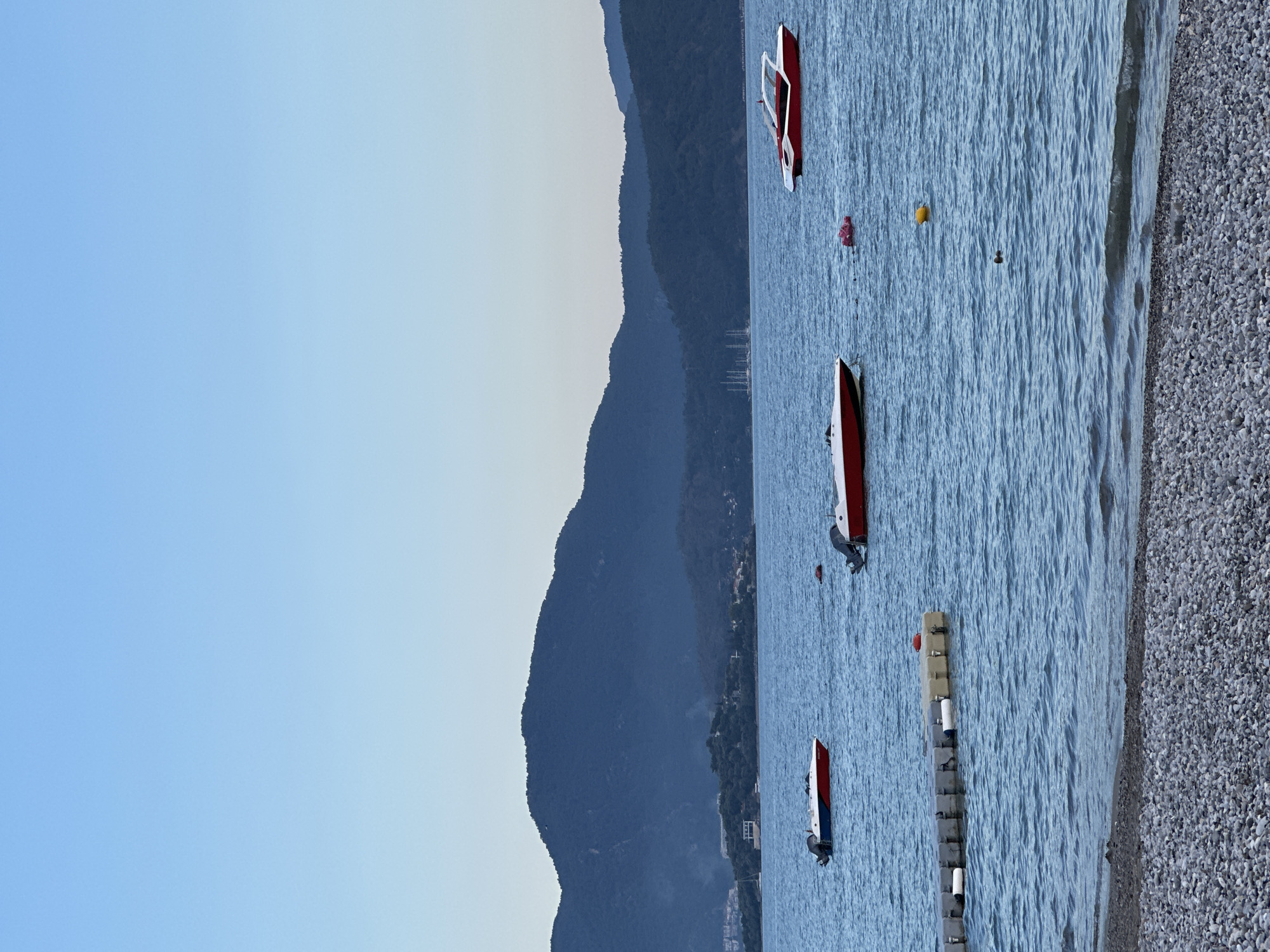 The image shows a coastal scene in Fethiye, Turkey. The water is a light blue color, and there are several small boats or jet skis docked in the water. There are also some buoys floating in the water. A rocky beach is visible on the right side of the image. In the background, there are dark blue mountains and a pale blue sky.