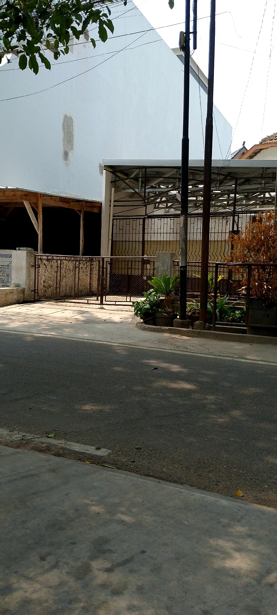 The image captures a street scene in Bandung City, Indonesia, on a sunny day. In the foreground, a gray asphalt road is visible with a concrete sidewalk to its left, shaded by dappled sunlight filtering through unseen trees. Beyond the sidewalk, a paved area leads to a property with a metal fence and a covered structure with a corrugated metal roof supported by a steel frame. A white wall forms the backdrop of the property, with a section showing signs of repair. Several thin metal poles, likely utility poles, are positioned in front of the property. A cluster of green plants is situated near the fence. The overall impression is a typical urban street view in a tropical climate.