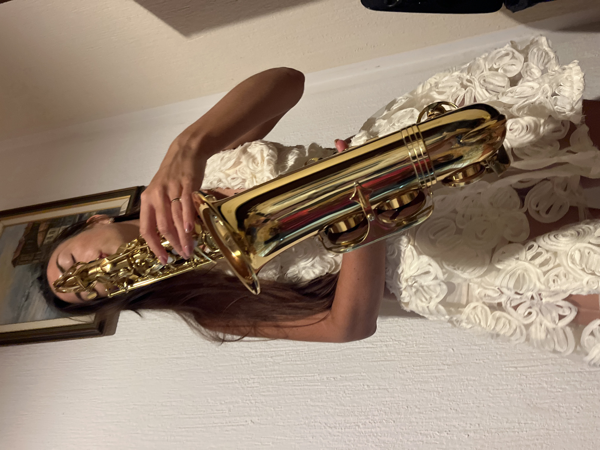 A woman in a white, rose-patterned dress holds a brass saxophone and is about to play it. She is standing indoors, in front of a white, textured wall. To the left, a framed painting hangs on the wall. To the right, a dark object, possibly a case, is visible. The lighting is dim, casting shadows on the wall. The scene appears to be in a room in Šušanj, Montenegro.
