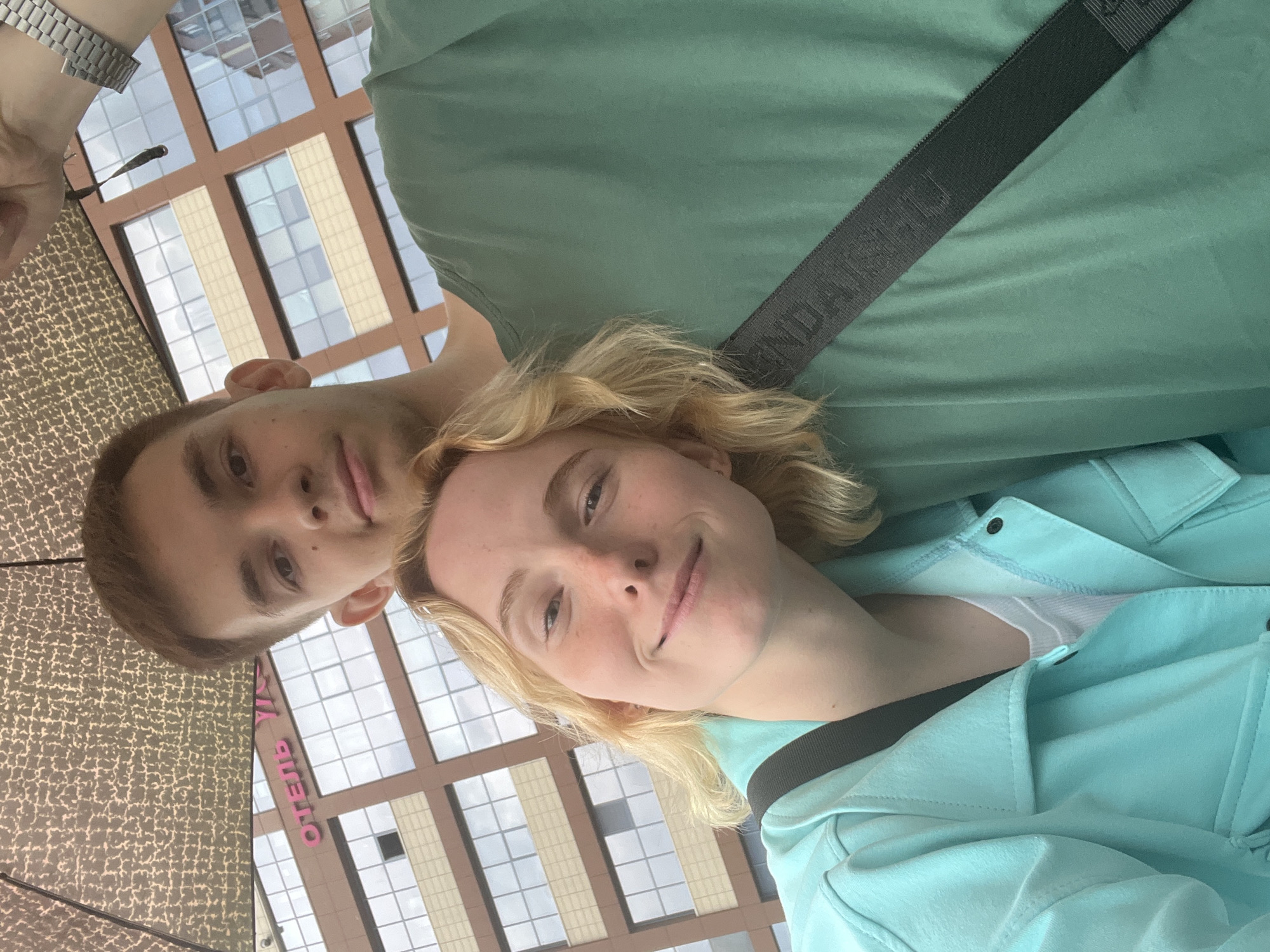This is a portrait-style photograph taken outdoors. The image features a young couple, a male and a female, close together. The male is on the left, his head turned towards the camera, showing his face and a portion of his upper body. He has short brown hair and is wearing a green t-shirt. The female is on the right, her face also turned towards the camera, smiling. She has blonde hair and is wearing a light blue jacket over a white top.
In the background, a modern building with large windows and a beige facade is visible. Part of a patterned surface, possibly an umbrella or canopy, is visible in the bottom left corner of the image. A dark strap with visible text is draped across the male's green shirt. The text reads "RUSSIA". Additionally, there is some pink text on the building in the background that appears to say "ОТЕЛЬ" which translates to "HOTEL" in Russian.
The lighting suggests it is daytime. The overall mood of the photo is casual and happy, with both individuals smiling. The location context indicates Tyumen, Russia.