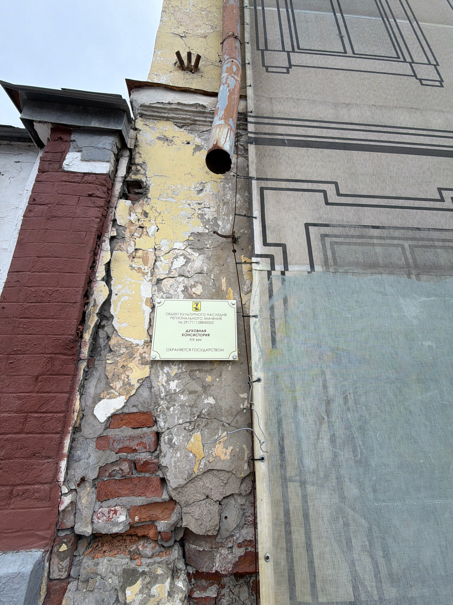 This is a close-up, low-angle photograph of the exterior wall of a building in Arkhangelsk, Russia. The wall is severely damaged, with large sections of plaster peeling away to reveal exposed brickwork. A rusty downspout runs vertically along the wall, showing signs of weathering and damage. A light green sign is affixed to the wall, bearing text in Cyrillic script, likely indicating historical or cultural significance. The sign is partially obscured by the damaged plaster. To the right of the damaged wall is a large, light-colored fabric stretched taut and secured with wires and grommets. This fabric appears to be a protective covering or a facade, with a dark, geometric pattern printed on it, resembling architectural elements. The overall impression is one of decay and ongoing preservation or renovation efforts. The image does not contain any people or explicit actions.