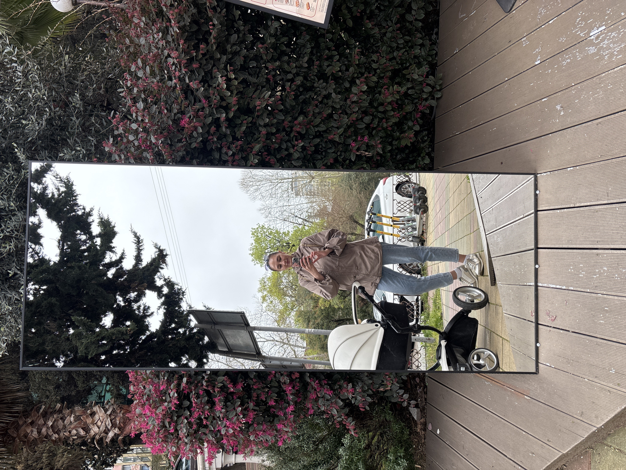 A woman stands near a baby stroller, reflected in a large rectangular mirror set outdoors in what appears to be a public space. She wears a tan jacket and light blue jeans, with silver sneakers. The stroller is white and black. In the reflection behind her are trees, a white van, and a row of scooters parked next to the van. One side of the mirror is positioned near lush greenery with pink flowers. The other side is next to a wooden deck-like platform. It is daytime. The location is likely in the city of Sochi, Russia.