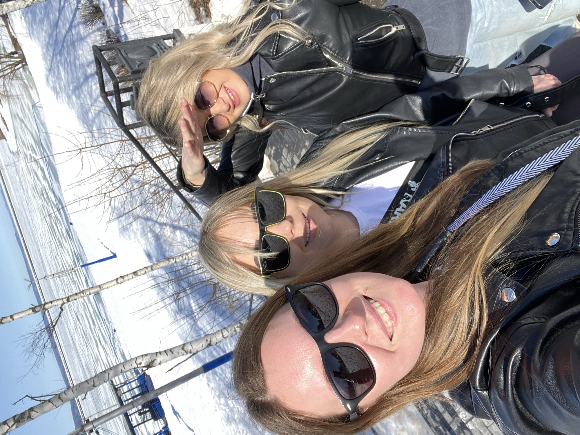 The image shows three women wearing sunglasses and black leather jackets. They appear to be taking a selfie, with the woman at the bottom of the frame holding the camera. The background shows a snowy landscape with birch trees, likely indicating a winter scene. The location is identified as the city of Chaikovsky, Russia. There is a bench partially visible on the left side of the image. The women are smiling, suggesting a positive and casual interaction.