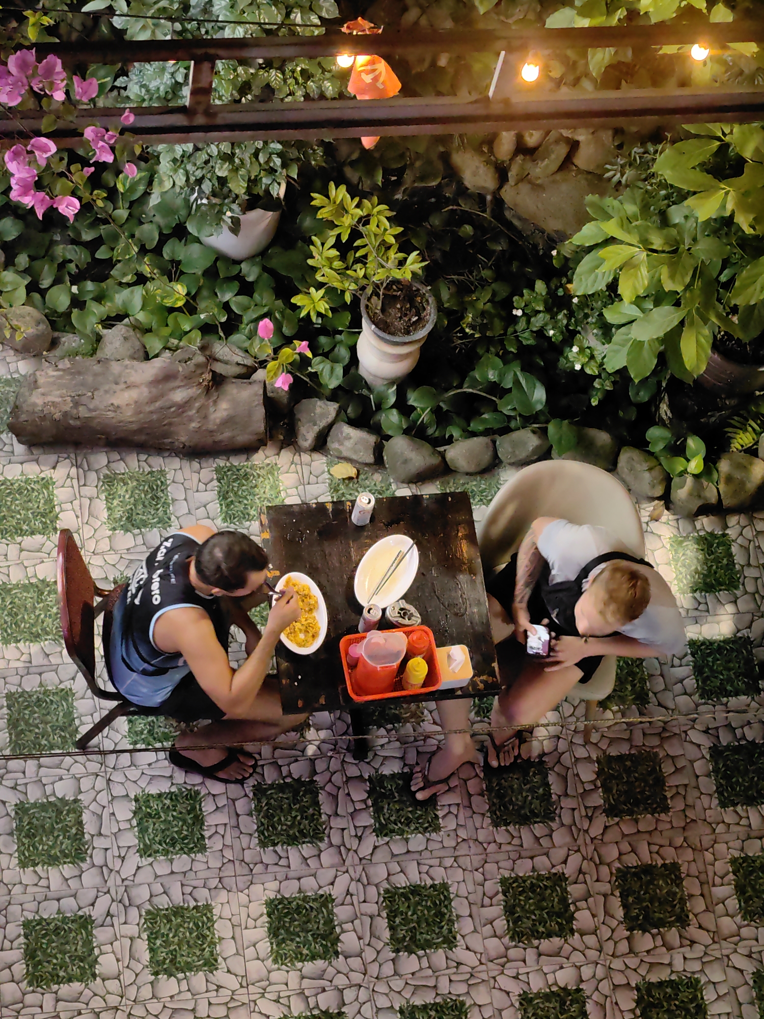 Aerial view of two individuals seated at a wooden table in an outdoor setting with tiled flooring and surrounding vegetation, identified as being in Bai Chay Ward, Vietnam. One person is actively eating a dish with chopsticks, while the other is looking at a mobile phone. A red tray containing condiments and a white plate with chopsticks rest on the table, along with what appear to be beverage cans. The flooring consists of tiles with embedded green patches. The vegetation includes flowering plants and various potted plants, with lighting fixtures visible overhead.