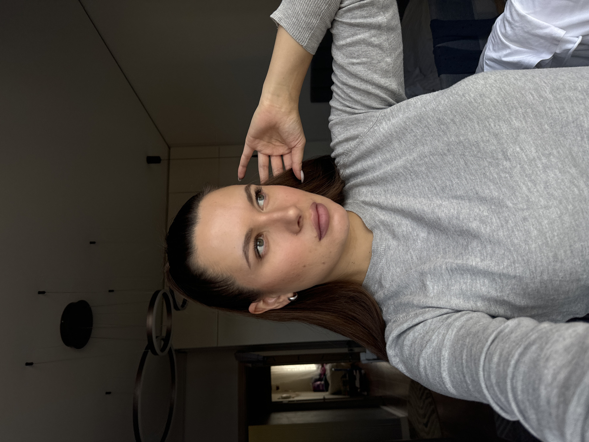 A young woman with brown hair, fair skin, and light-colored eyes is featured in a portrait-style indoor image. She is seen from the chest up, with her head slightly tilted and her gaze directed upwards and to her left. Her left hand is raised, with her fingers gently resting near her temple, while her right arm is bent. She is wearing a simple, light grey, long-sleeved ribbed top or sweater and has a small, minimalist hoop earring in her left ear. Her lips are full and have a subtle pink tint.
The background suggests a modern, minimalist interior, likely a bedroom or living area. To the left of her head, against a dark wall, a distinctive light fixture or art piece is visible, consisting of a dark circular base from which thin wires extend, possibly holding small lights. Behind her, elements of a bed or sofa are visible, including white and darker fabrics. A reflection in the deeper background hints at another part of the room. The lighting appears subdued, contributing to a calm and relaxed atmosphere. The woman's expression is serene, as if posing casually for the photograph. No text is visible in the image. The location context for this image is Akulovo (Gp Kubinka), Russia.