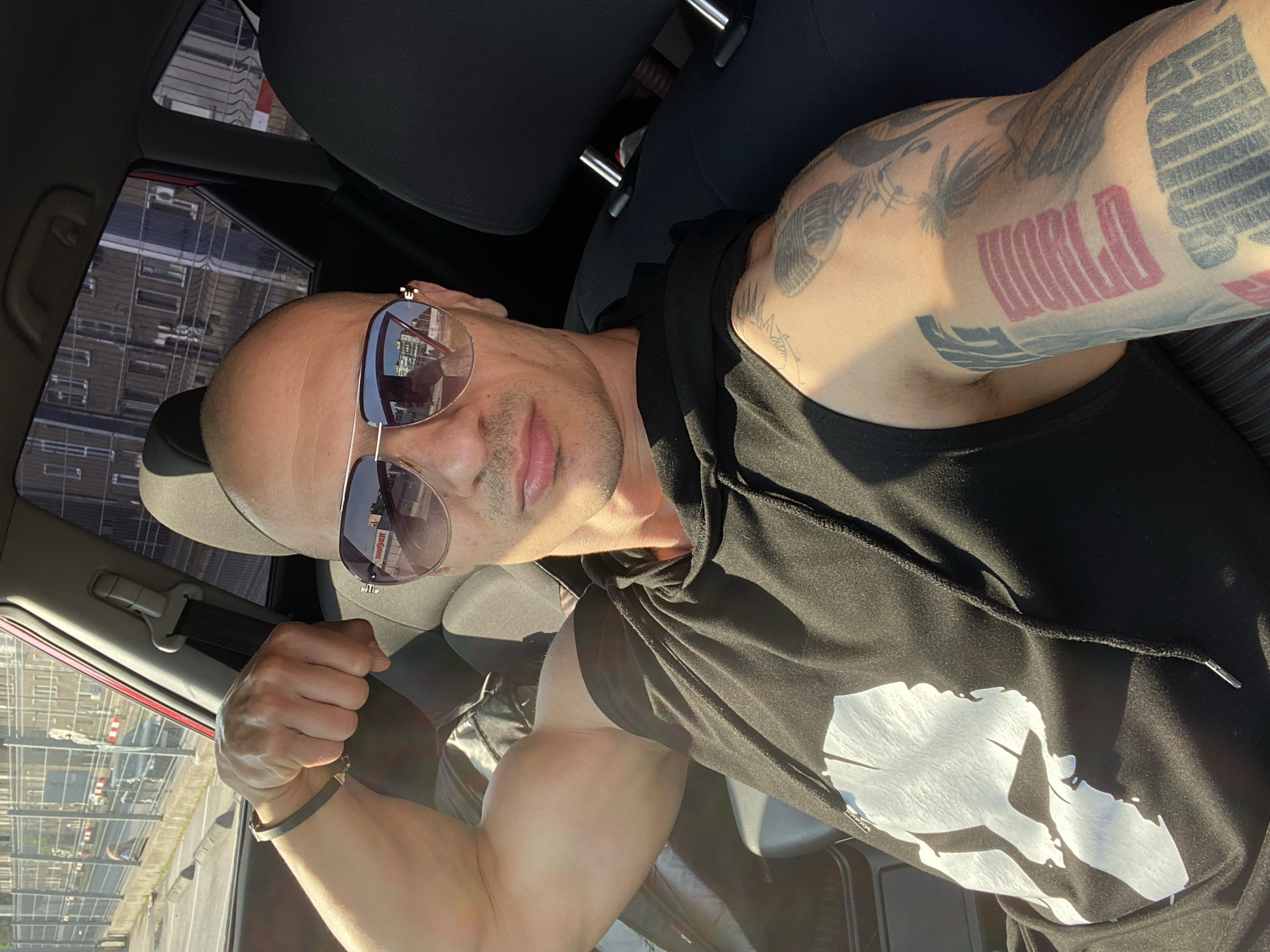 A bald man wearing mirrored aviator sunglasses and a black sleeveless hoodie is captured in a selfie taken inside a car, likely from the driver's seat. He has extensive tattoos on both arms, with the word "WORLD" prominently visible in red block letters on his left bicep (visible on the right of the image). He is flexing his right arm, showcasing his bicep, and a black bracelet is visible on his left wrist. The black hoodie features a large white, abstract graphic on the front, resembling a stylized face or landmass.
Through the car window and reflected in his sunglasses, an urban landscape with multiple buildings, possibly apartment blocks or commercial structures, and a parking lot or construction area is visible. The strong sunlight and cast shadows indicate it is daytime, possibly morning or late afternoon. The man has a neutral expression, conveying confidence. The setting is an urban environment, with the specific location being Pleven, Bulgaria.