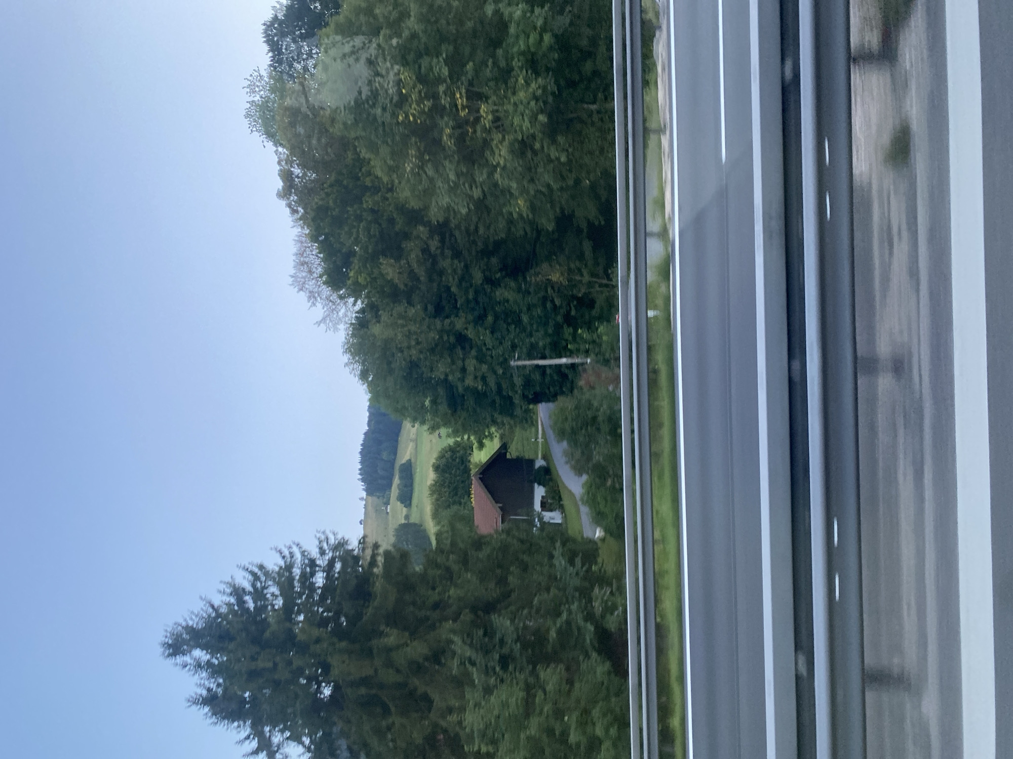 The image shows a view from a highway in Anger, Germany, looking towards a rural landscape. The foreground features highway guardrails and a portion of the road. Beyond the highway, a dark-colored house with a red-tiled roof is visible, nestled amongst lush green trees. A winding road leads up to the house. In the background, rolling green hills are dotted with trees and a small white structure, possibly a chapel or a small building, on a distant hilltop. The sky is a pale blue, suggesting it is either early morning or late evening. The overall scene depicts a tranquil, natural environment characteristic of the Bavarian countryside.