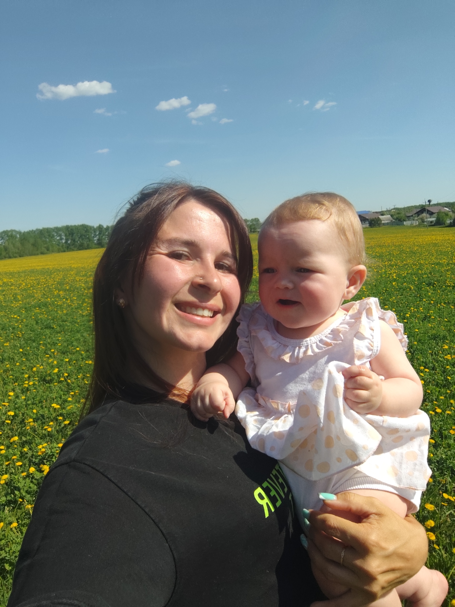 A woman holds a baby in a field of yellow flowers in Фоки, Russia. The woman is wearing a black t-shirt and has long, dark brown hair and fair skin. She is smiling and looking at the camera. The baby is wearing a white dress with a light-colored polka-dot pattern and has light blonde hair and fair skin. The background is a blue sky with a few white clouds. There is a line of trees and some houses in the distance.