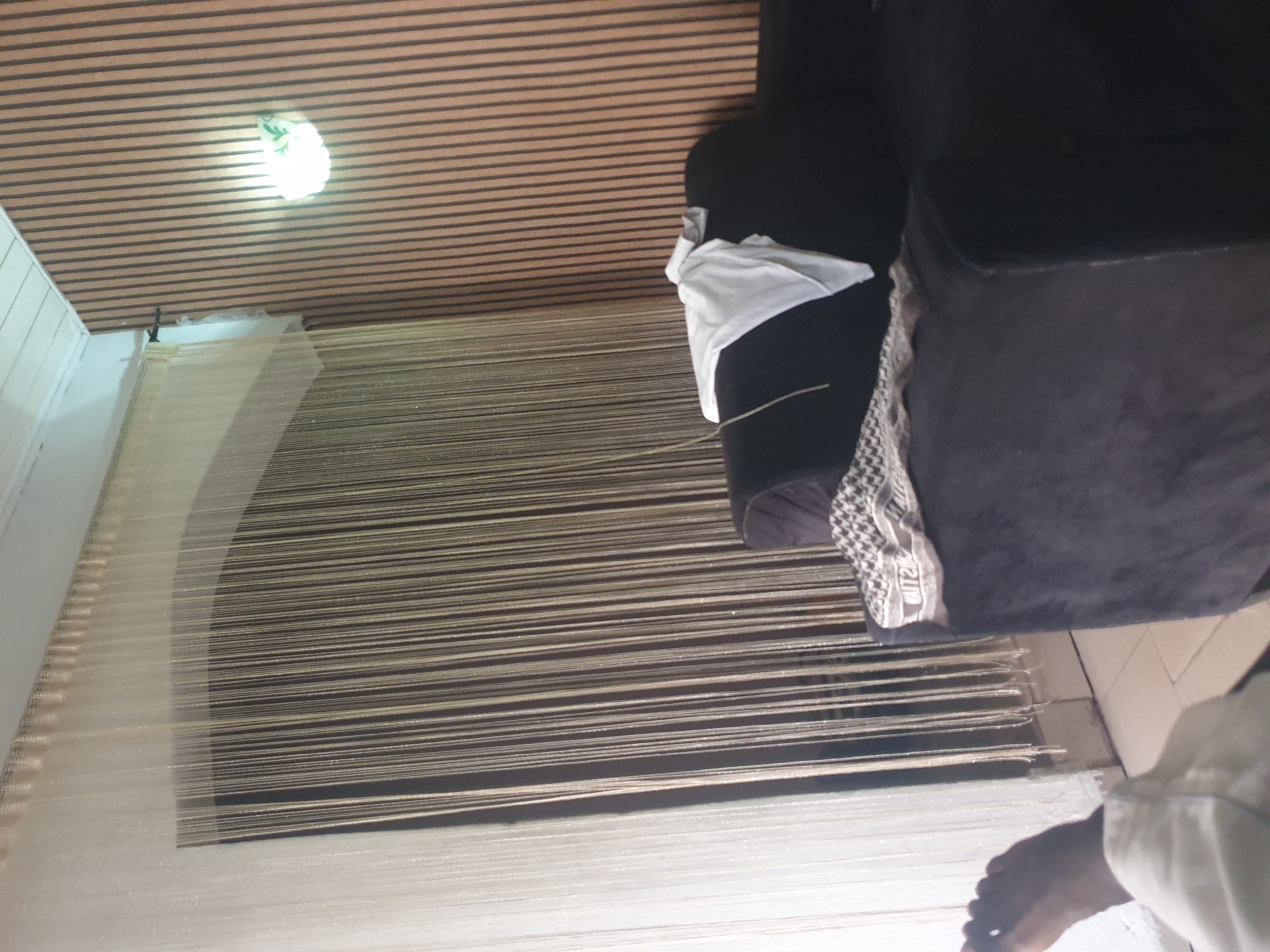 The image depicts an interior scene of a room, possibly a living area. A dark-colored sofa is positioned in the foreground, partially draped with a white cloth. A patterned textile, likely a scarf or throw, is placed on the left armrest of the sofa. To the left of the sofa, a person's bare foot and lower leg are visible, clad in white fabric. Behind the sofa, a wall with vertical wooden slats is illuminated by a wall-mounted light fixture. To the left of the slatted wall, a door or window is covered by a sheer, beaded curtain, creating a textured backdrop. The ceiling is white and appears to be paneled. The overall impression is one of a casually arranged domestic space.