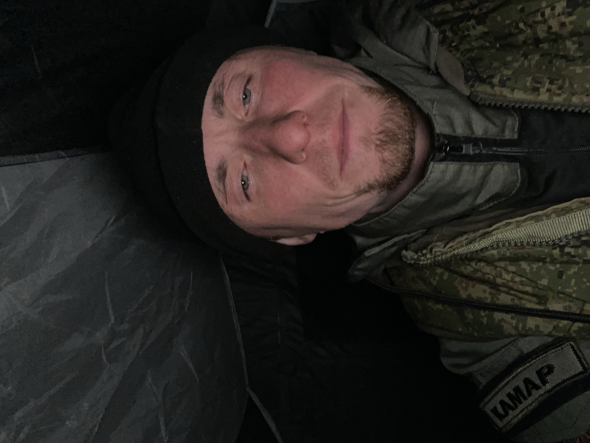 A man wearing a black balaclava, a camouflage jacket, and a patch that reads "KAMAP" is lying down. The environment appears dark and confined, possibly inside a tent or enclosed space. The image is taken from a high angle, looking down at the man's face. The location is unknown and not determinable from the image itself.