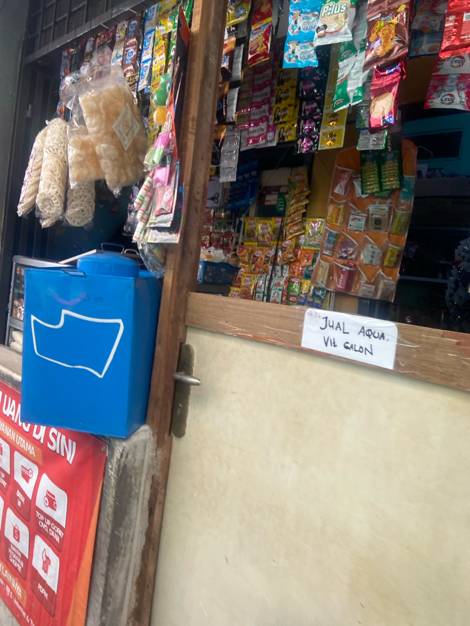 The image depicts a small roadside store in North Jakarta, Indonesia. The store appears to be a kiosk or small shop, characterized by a wooden frame and shelves filled with various packaged goods. Hanging from the ceiling are numerous colorful packets of snacks and other small retail items. Prominently displayed is a large blue container, possibly a water cooler or storage bin, with a white outline of a stylized shape on its front. To the right of the blue container, a wooden plank holds a white sign with handwritten text in Indonesian that reads "JUAL AQUA. VIL GALON." which translates to "Selling Aqua. Via Gallon." This suggests the store sells bottled water.
Below the blue container, a red banner with white text and icons is partially visible, likely advertising services or products. The overall environment suggests a typical street-side vendor setup in a busy urban area. The lighting indicates it is daytime. No people are visible in the image, and the focus is on the merchandise and the storefront structure.