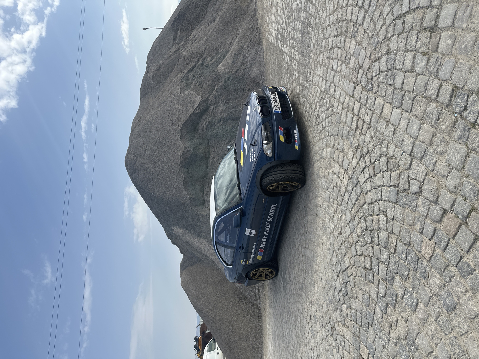 The image depicts a blue BMW rally car parked on a cobblestone road in Negovan, Bulgaria. The car has "JEKOV RALLY SCHOOL" written on its side and a Bulgarian license plate "CB 3446 OB" on the front. Behind the car is a large gray rocky hill. Above the hill is a blue sky with a few thin clouds and some electrical wires. A white van and other miscellaneous items are partially visible at the bottom left of the image.