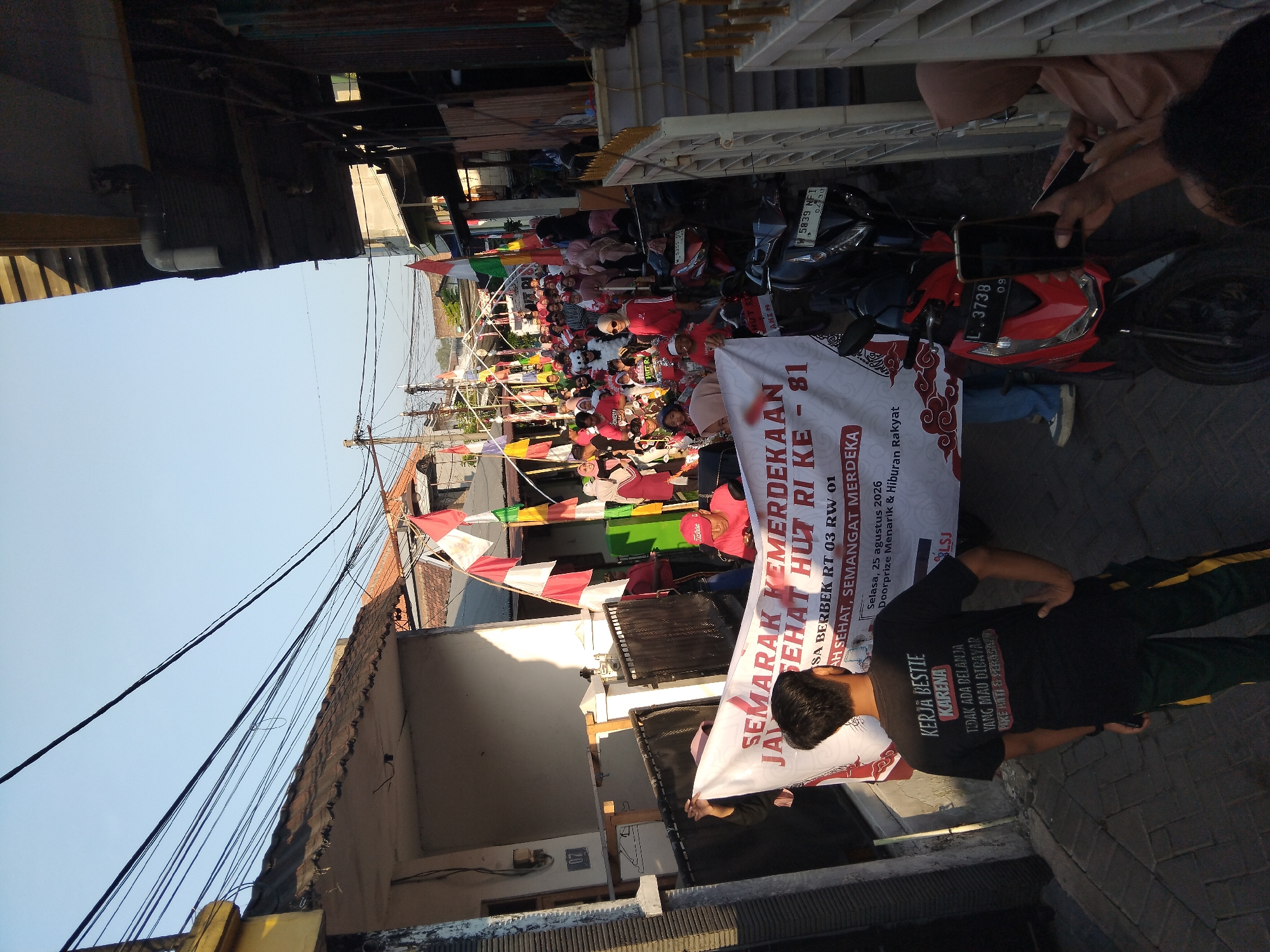 The image depicts a street scene in Kepuh Kiriman, Indonesia, possibly during a celebratory event. A group of people, including men, women, and children, are gathered in a narrow street adorned with numerous Indonesian flags and pennants strung across overhead wires. The people are mostly wearing red and white attire, suggesting a patriotic theme. In the foreground, two individuals are holding a large banner with red text that reads "SEMARAK KEMERDEKAAN HUT RI KE - 81" and further text relating to a specific location and date. Several motorcycles are parked along the sides of the street. Buildings with tiled roofs and balconies line the street, with overhead electrical wires creating a dense network above. The lighting suggests it is either early morning or late afternoon, with a clear sky.