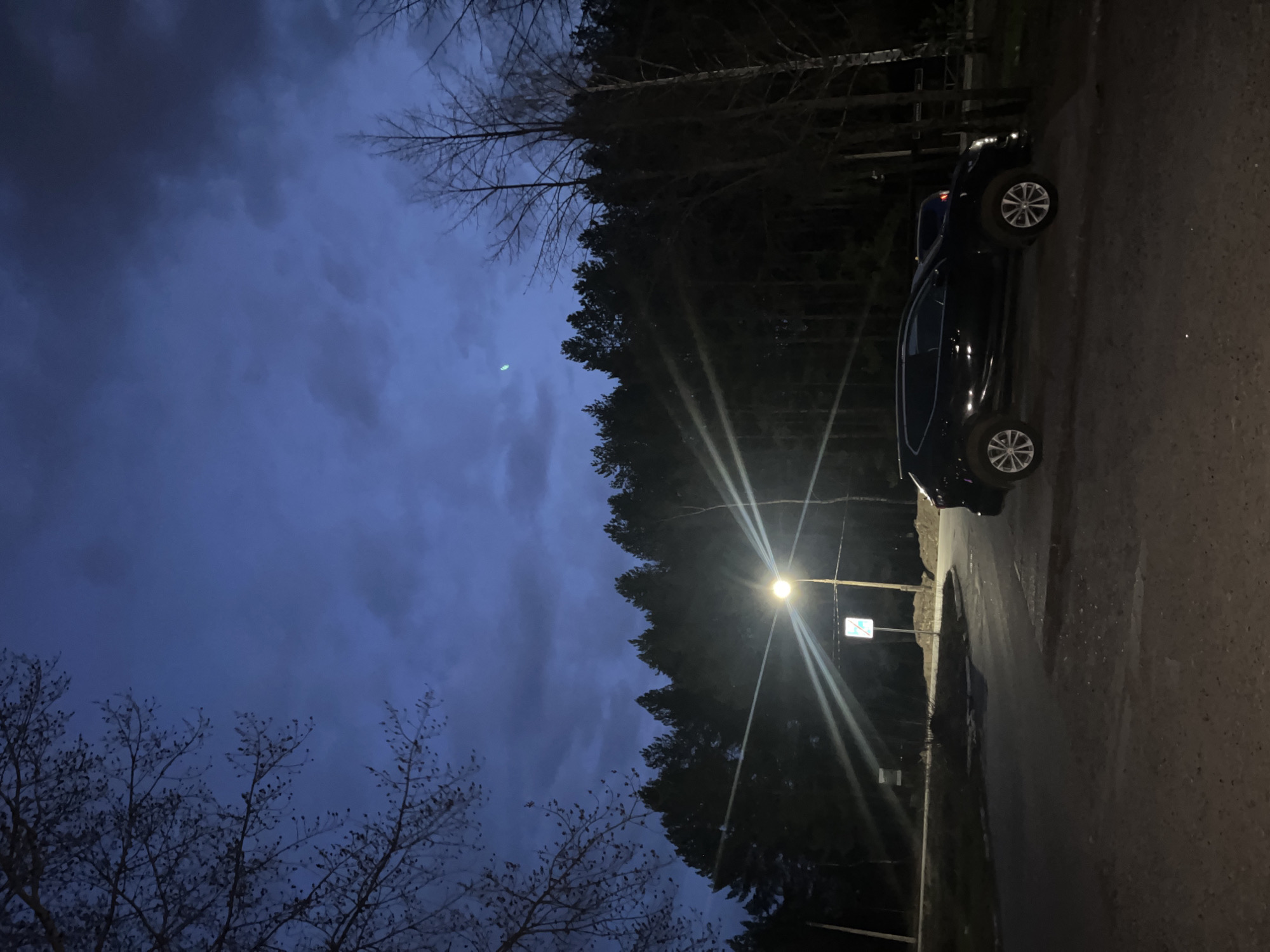 The image presents a dark outdoor scene in what appears to be a residential or semi-rural area near the city of Chaikovsky, Russia. A dark-colored car is parked on the side of a paved road. The sky is dark blue with scattered clouds, and there are trees bordering the road, some of which are bare, indicating it might be late autumn or early spring. A street light illuminates a small area of the road and a road sign. The general atmosphere suggests it is nighttime.