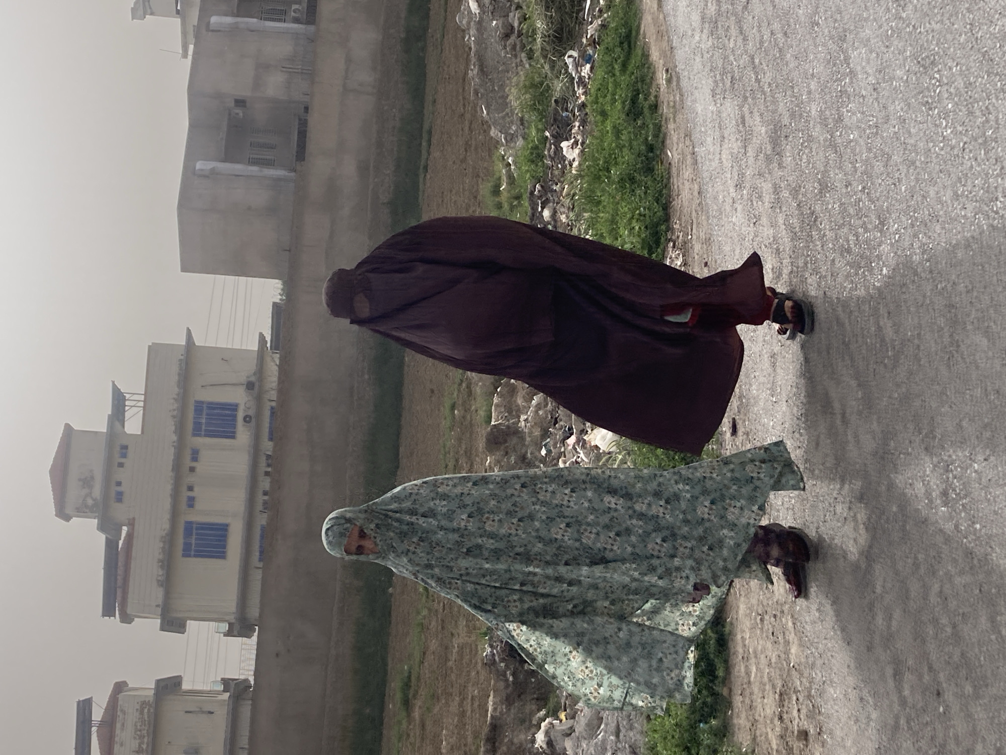Two women wearing burqas walk along a road in Quetta City Tehsil, Pakistan. One woman wears a dark brown burqa and the other wears a burqa with a pale green floral pattern. Buildings line the street on one side. Vegetation and refuse are visible on the other side of the road. The day appears overcast.