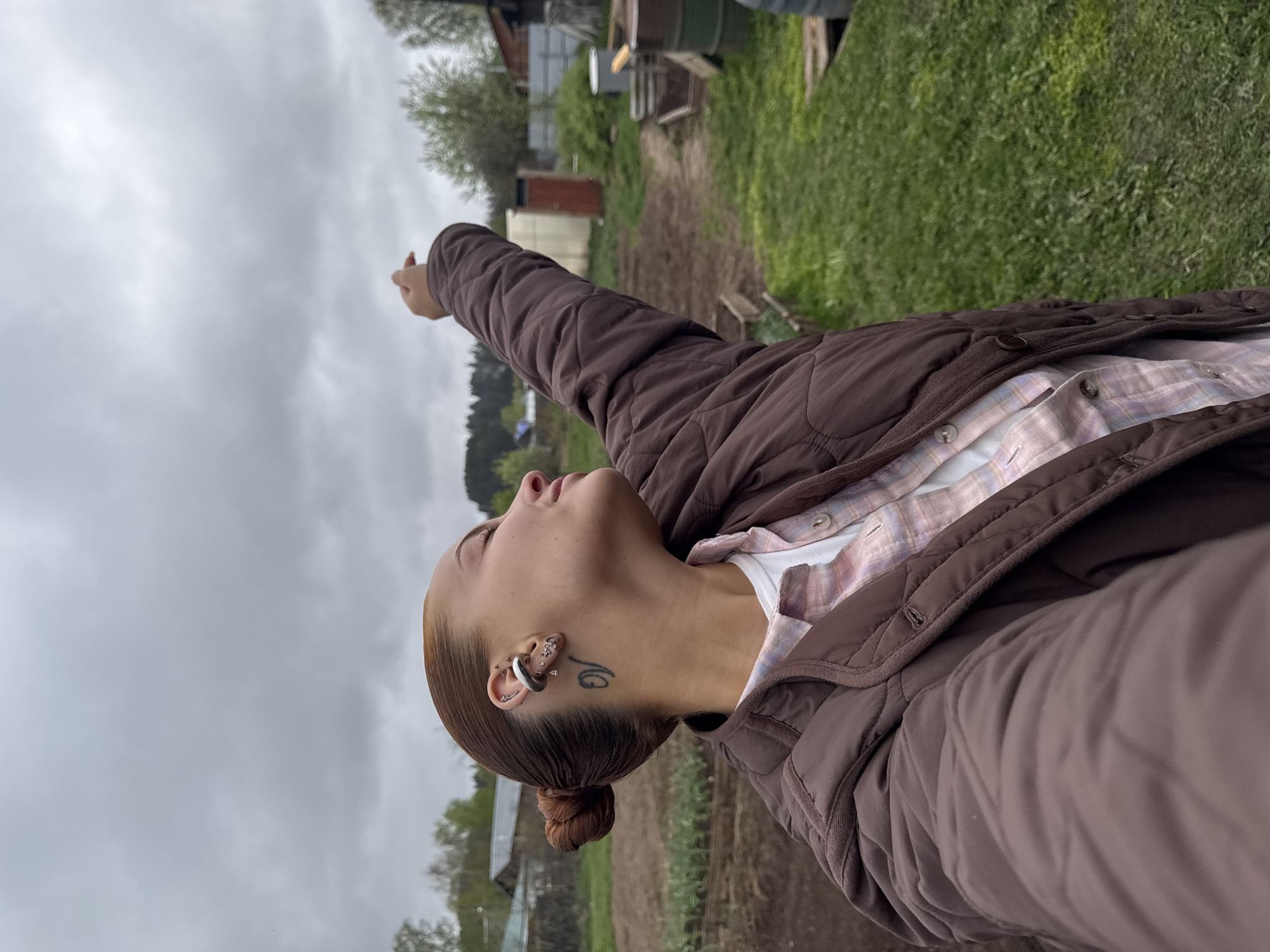 A young woman with fair skin and light brown hair pulled back into a bun is the central subject of the image. She is wearing a brown quilted jacket over a light pink and white plaid shirt. Her left arm is extended upwards, pointing towards the sky. Her head is tilted back as she looks towards the same direction. Several ear piercings and a tattoo near her left ear are visible.
The background shows a rural or semi-rural landscape under an overcast sky. A mixture of green grass and plowed brown soil indicates a garden or farm area. There are a few buildings in the distance, including what appears to be a barn or shed. The location is Chastyye, Russia.