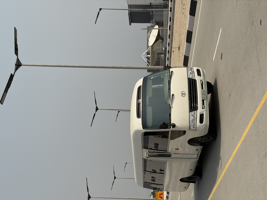 The scene depicts a white Toyota Coaster minibus in motion on a paved road in Alaguntan, Nigeria, under a hazy sky. The vehicle, bearing Nigerian license plate "ARO-422AA," is angled towards the viewer's right, occupying the left lane marked with a solid yellow line. Multiple tall, modern streetlights with Y-shaped LED fixtures line the road. To the right of the road, a median strip with a black and white striped curb separates the roadway from a sparsely vegetated area. In the background on the right, a large white satellite dish is visible near a building with a complex white pyramidal roof structure and another modern building with a dark facade. On the left side of the road, a yellow triangular warning sign with "HITECH" text is partially visible. No individuals are discernible within or around the minibus.