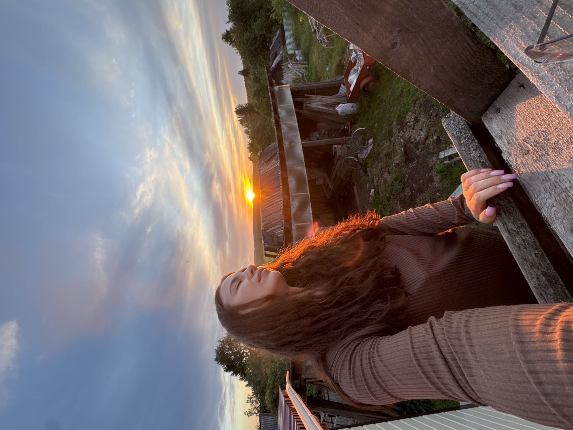 A woman stands on a wooden structure in Nizhnyaya Gar', Russia. The sunset casts a warm glow on her face and hair. Behind her, a cluttered rural landscape includes a dilapidated building, vegetation, and various objects, including a red cart and a bicycle. The sky is filled with streaks of light-colored clouds.